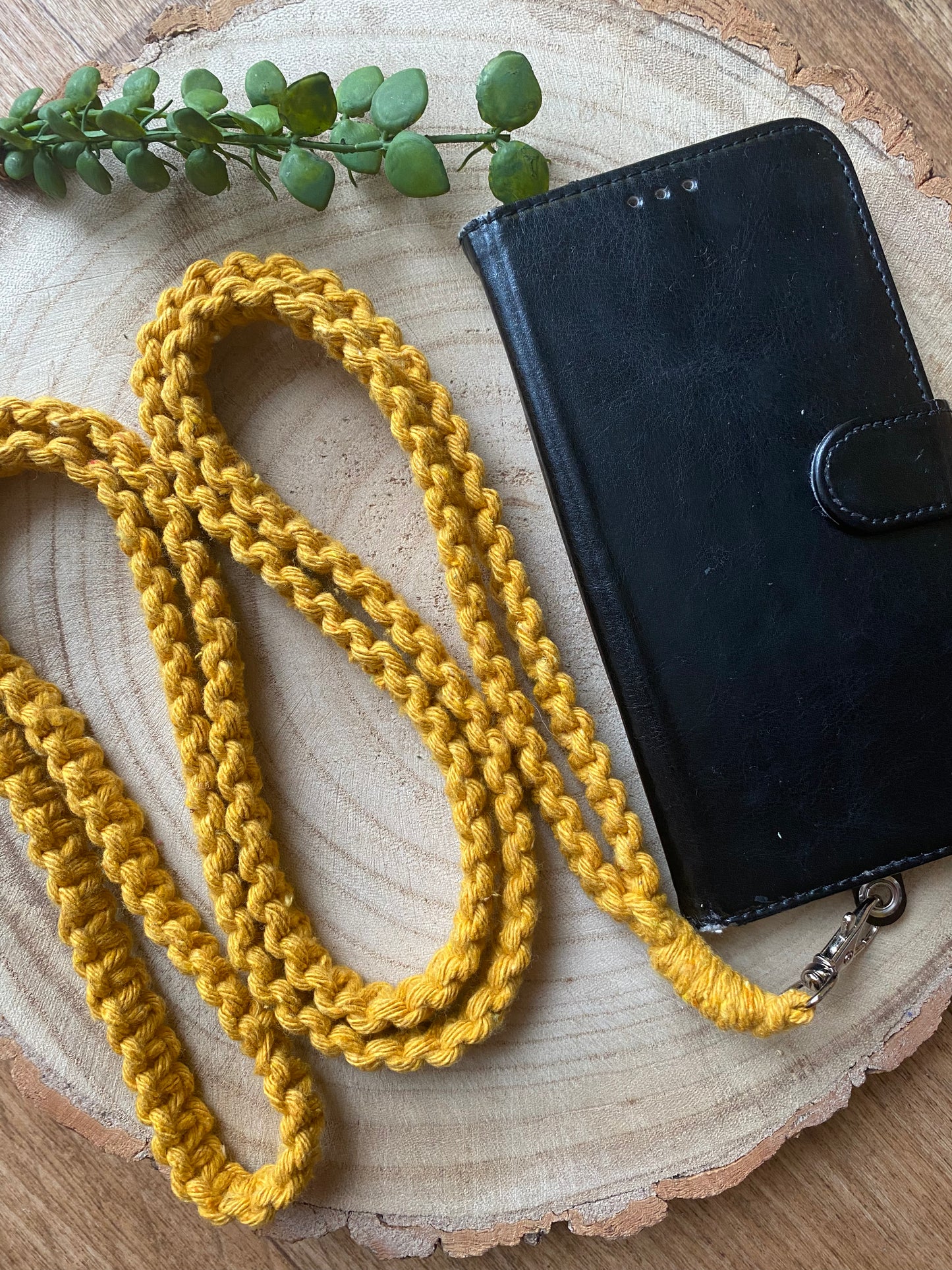A long, mustard-yellow braided rope with a metal clasp is coiled on a wooden surface next to a black leather phone case. This eco-friendly setting features a handmade Recycled cotton crossbody phone strap from Macra-Made-With-Love. A small sprig of green eucalyptus leaves lies on the edge of the wooden surface.