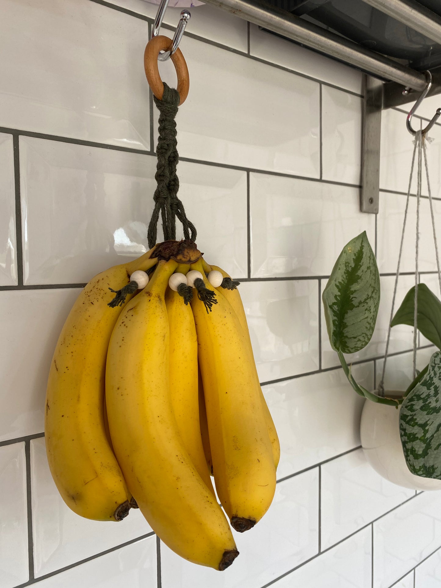 A bunch of ripe yellow bananas hangs from a Macra-Made-With-Love Banana Hanger on a wooden hook attached to a white tiled wall. Nearby, an eco-friendly home decor potted plant with green leaves is suspended in a white hanging planter.
