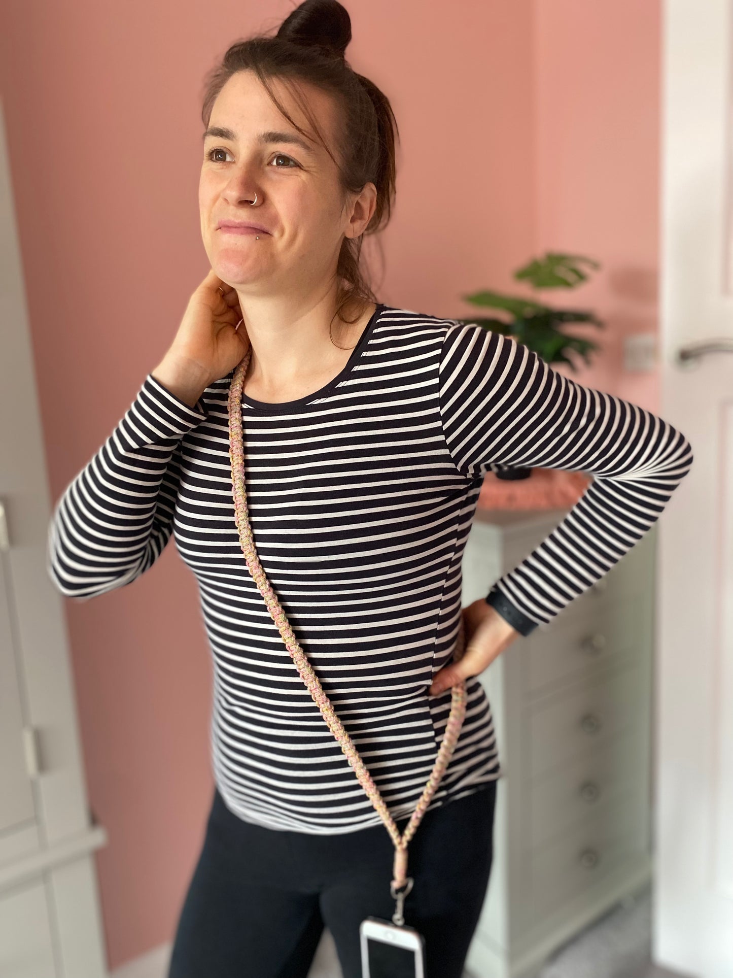 A woman stands in a room with her hand on her neck, looking to the side and slightly smiling. She is wearing a black and white striped shirt and black pants. A phone attached to a Macra-Made-With-Love Recycled cotton crossbody phone strap hangs around her neck. The background includes pink walls and some furniture.