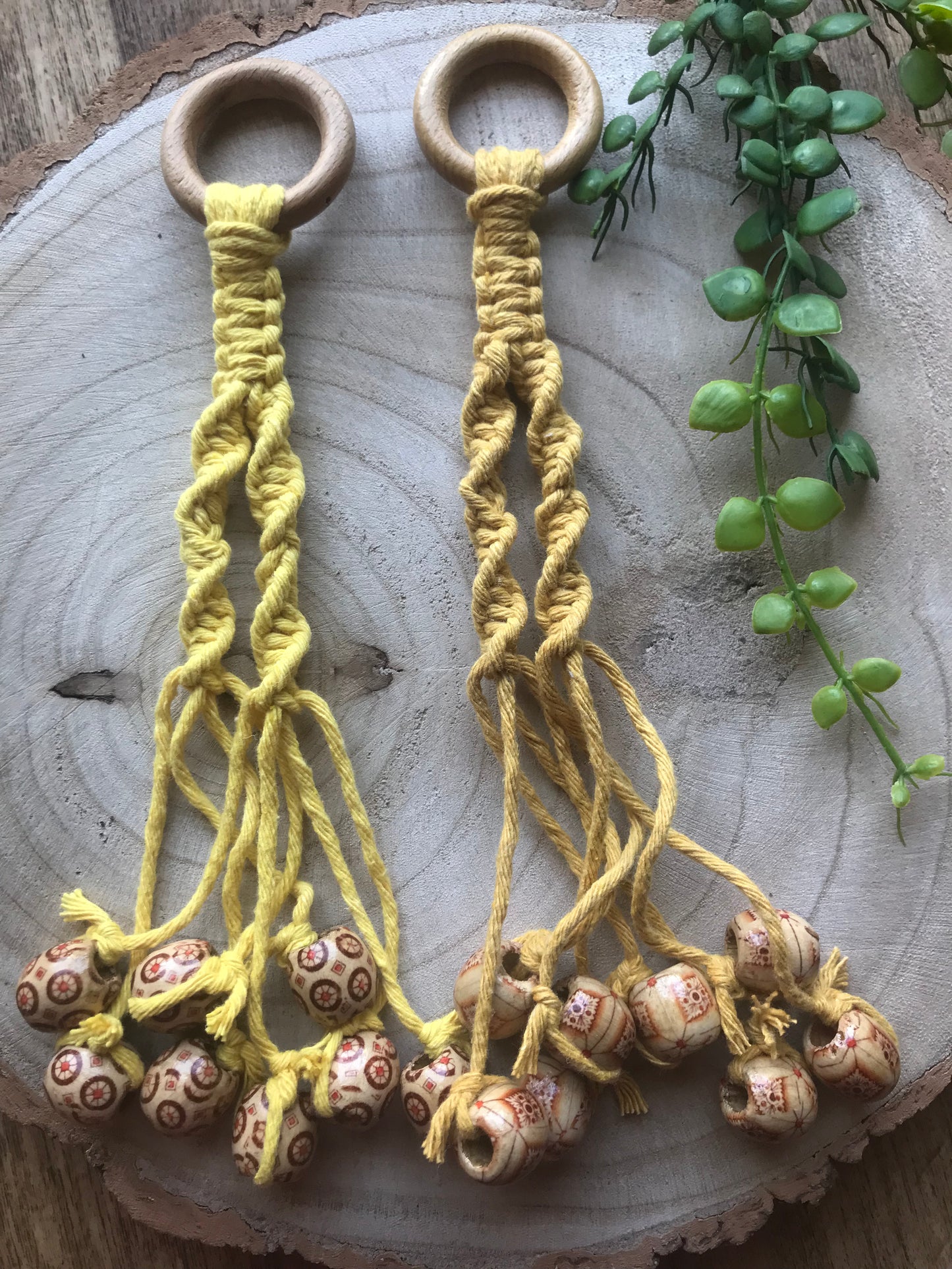 Two Banana hangers with wooden rings and woven yellow cord are laid out on a wooden circular surface. Each Banana hanger ends with strings adorned with patterned wooden beads. An eco-friendly home decor piece by Macra-Made-With-Love, there is a green plant with round leaves to the right of the Banana hangers in the image.