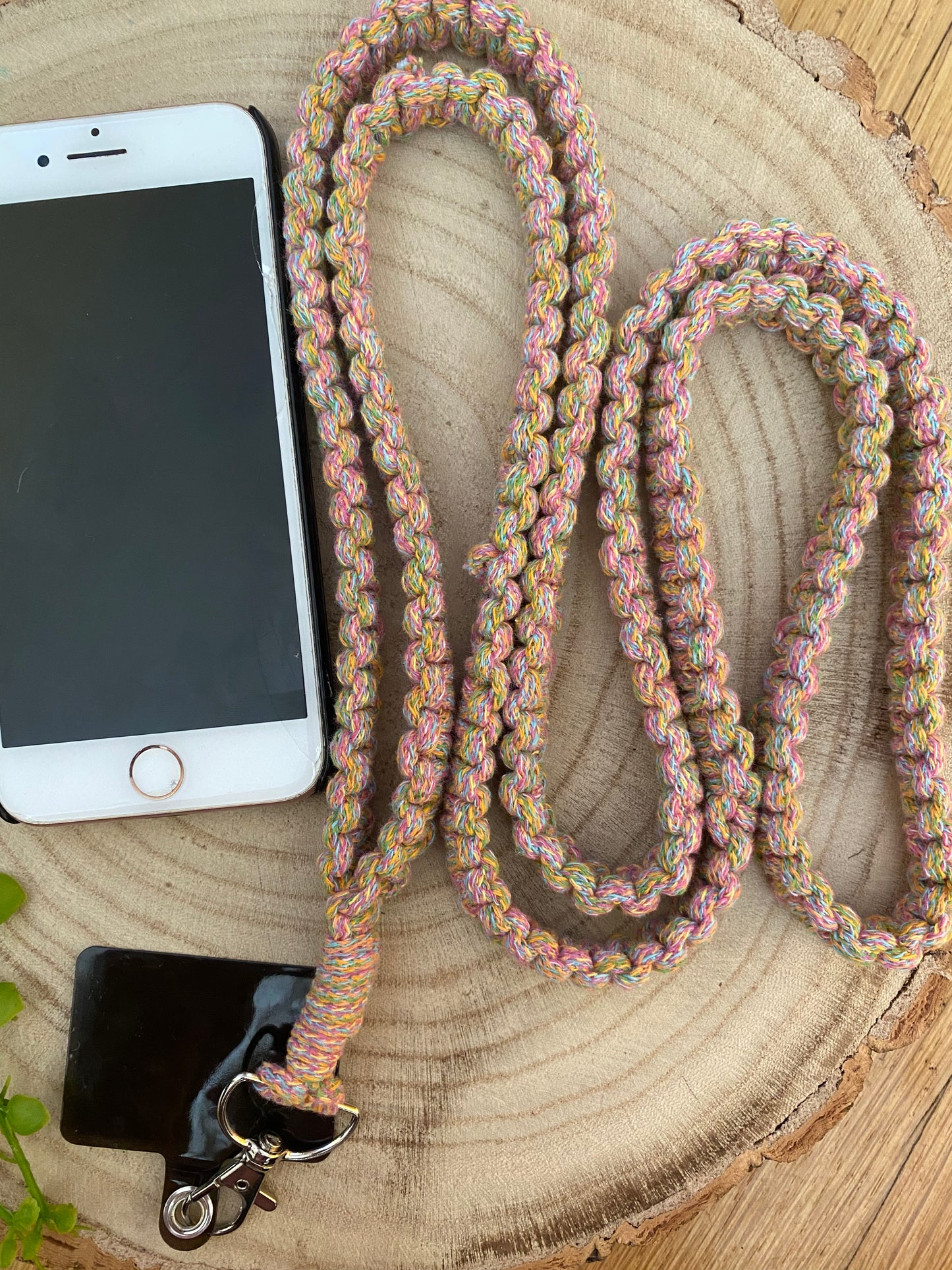 A smartphone with a white front and gold-colored home button rests on a wooden surface. It is attached to an eco-friendly, colorful, braided lanyard with a metal clip. The handmade Macra-Made-With-Love Recycled cotton crossbody phone strap loops around the phone and spreads out on the wood. A small plant is partially visible.