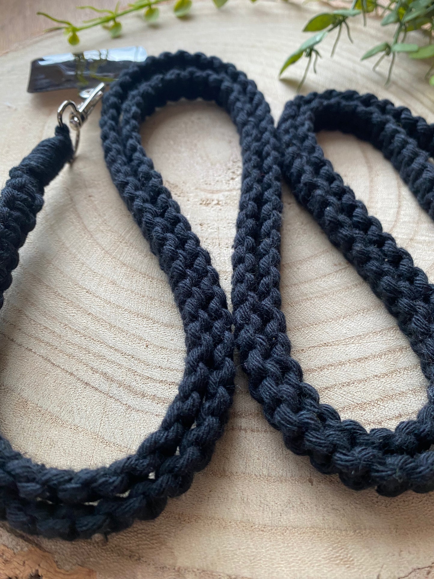 A black braided leash with a metal clasp is coiled smoothly on a wooden surface. Greenery is positioned around the edges, framing this Recycled cotton crossbody phone strap by Macra-Made-With-Love with a natural backdrop.