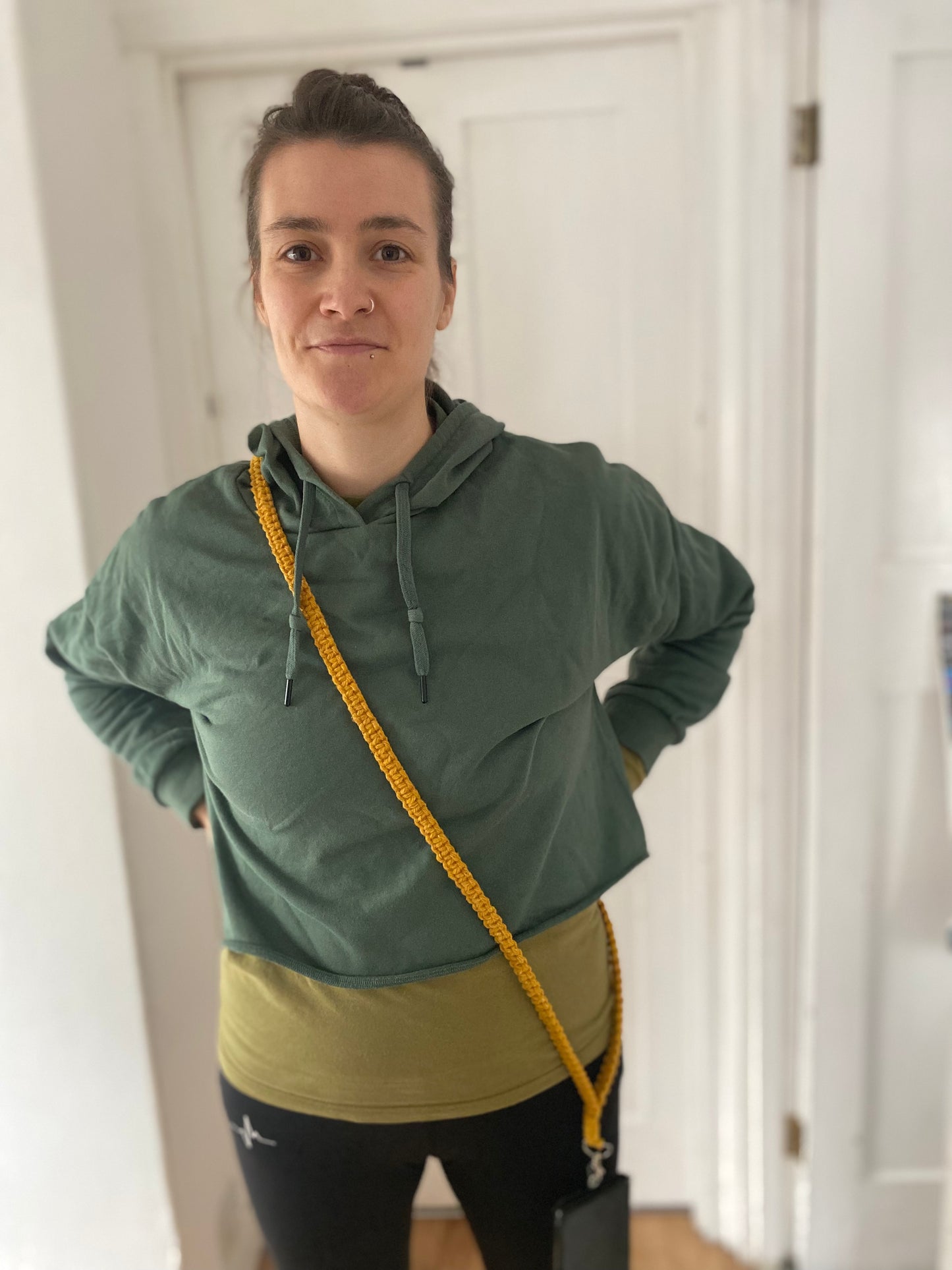 A person with long hair tied back stands indoors against a white wall, wearing a green hoodie with a yellowish-green section at the bottom, black leggings, and a Recycled cotton crossbody phone strap by Macra-Made-With-Love diagonally across their body. They have their hands on their hips and are looking at the camera.