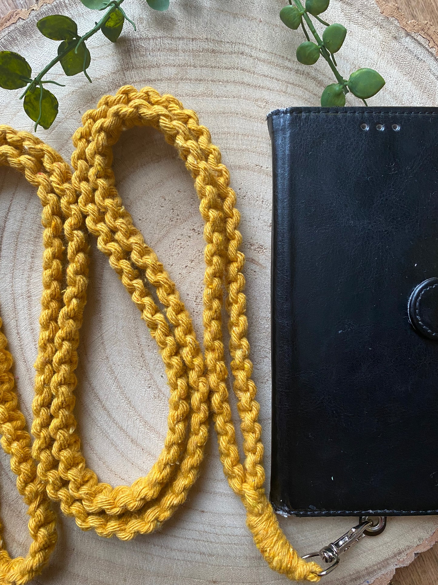 A black leather wallet with a button clasp lies open on a wooden surface next to an eco-friendly, mustard yellow, handmade Recycled Cotton Crossbody Phone Strap from Macra-Made-With-Love. Two green vine branches are seen at the top. The wooden surface has concentric rings, indicating it is a cross-section of a tree trunk.