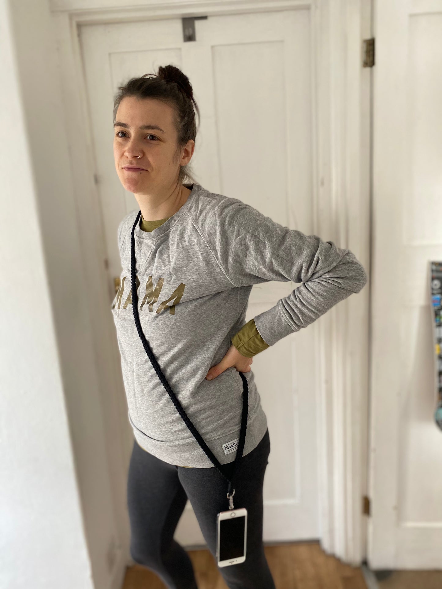 A woman stands indoors, slightly leaning to one side with a hand resting on her hip. She wears a gray long-sleeve shirt with "MAMA" printed on it and black leggings. A Recycled cotton crossbody phone strap by Macra-Made-With-Love, an eco-friendly phone accessory, hangs across her body. The background features white doors.