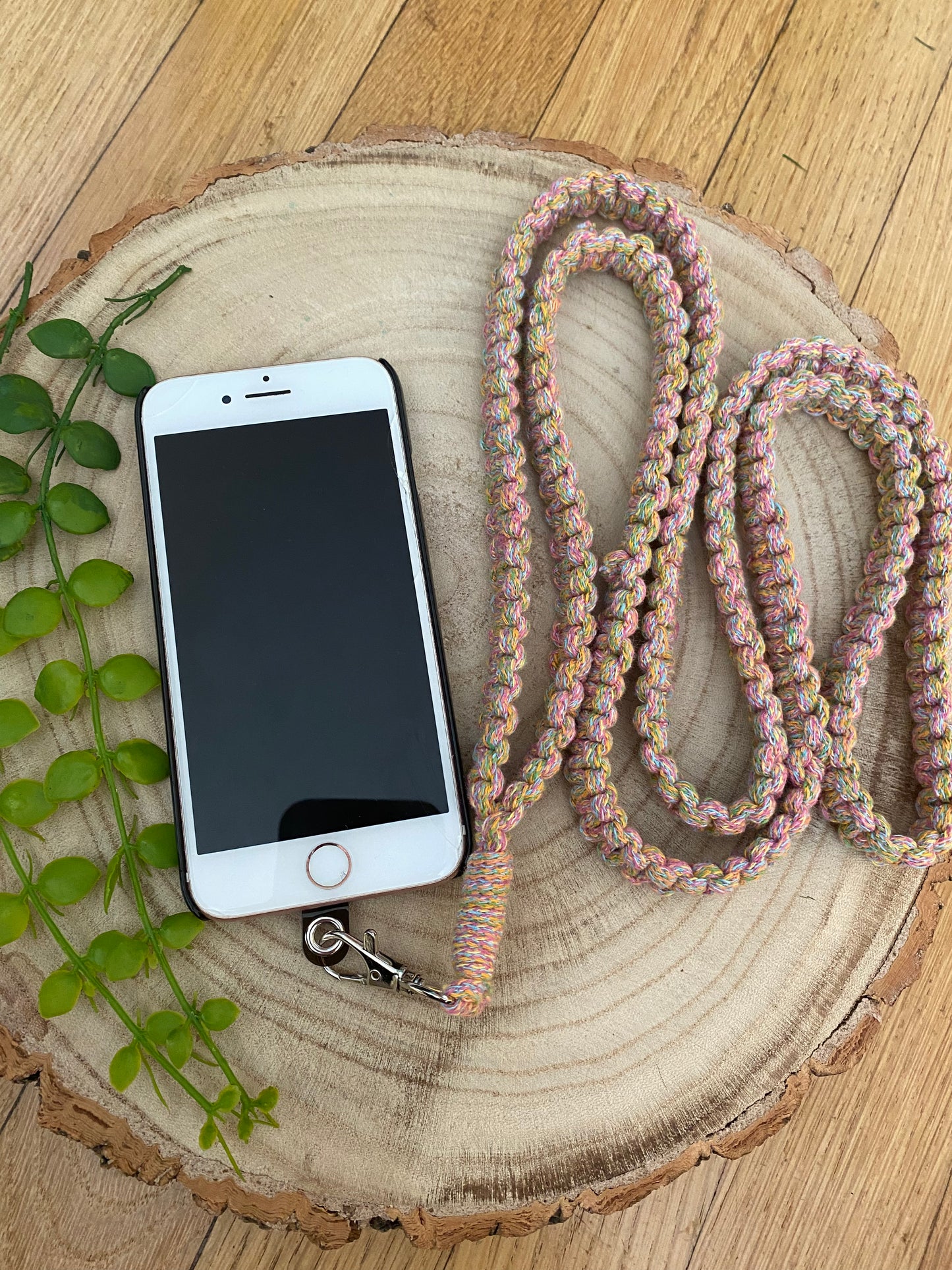 A white smartphone lies on a circular wooden slice beside a trailing green plant. Attached to the phone is a multicolored, braided lanyard with a metal clasp, coiled in a loose loop. This Recycled cotton crossbody phone strap by Macra-Made-With-Love adds an eco-friendly touch to the phone accessory. The background is a wooden surface.