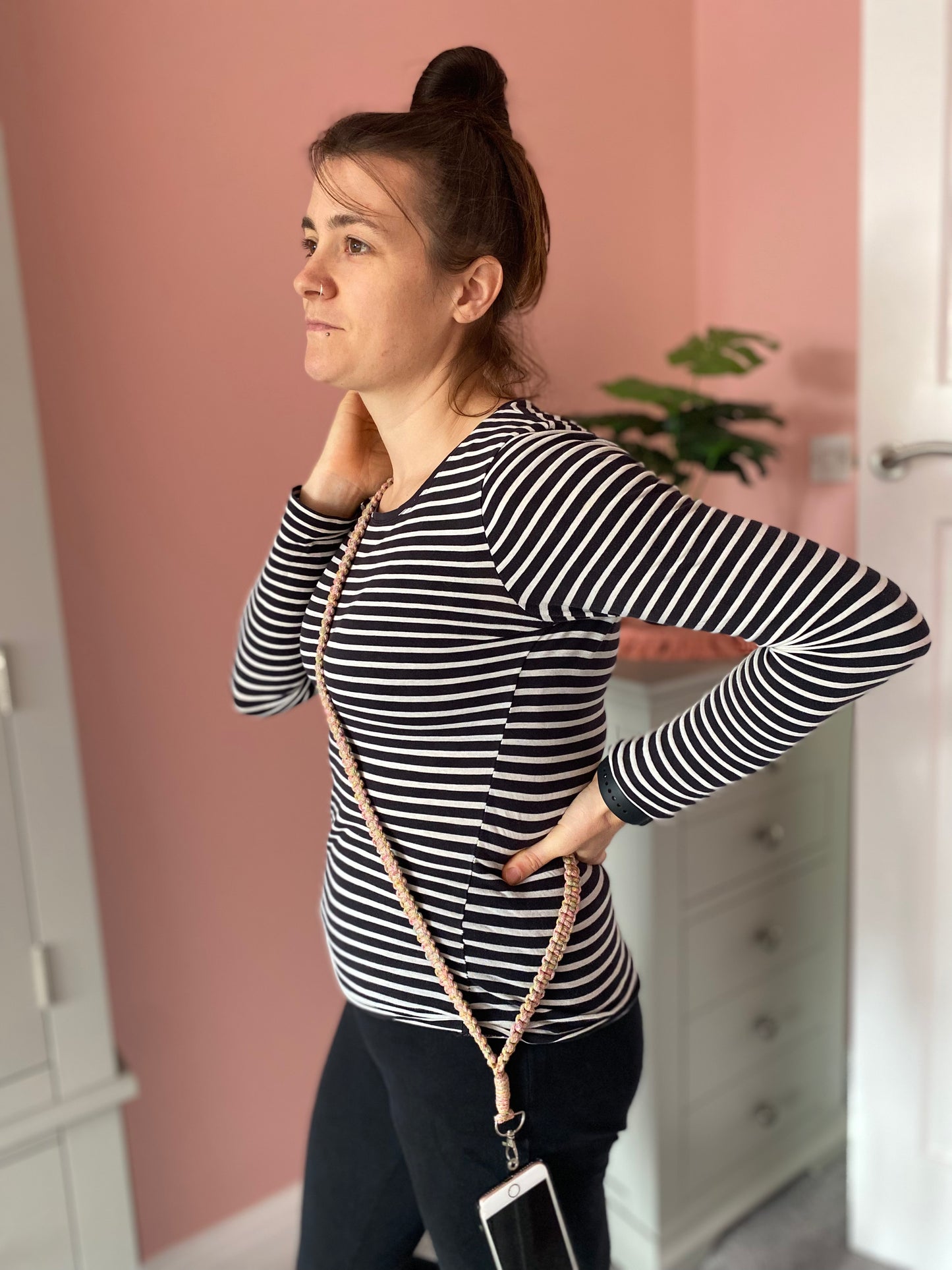 A woman stands in a room with a pink wall, wearing a black-and-white striped long-sleeve shirt and black pants. She has a Macra-Made-With-Love recycled cotton crossbody phone strap draped across her body and holds her right hand to her neck. A plant and a cabinet are visible in the background.