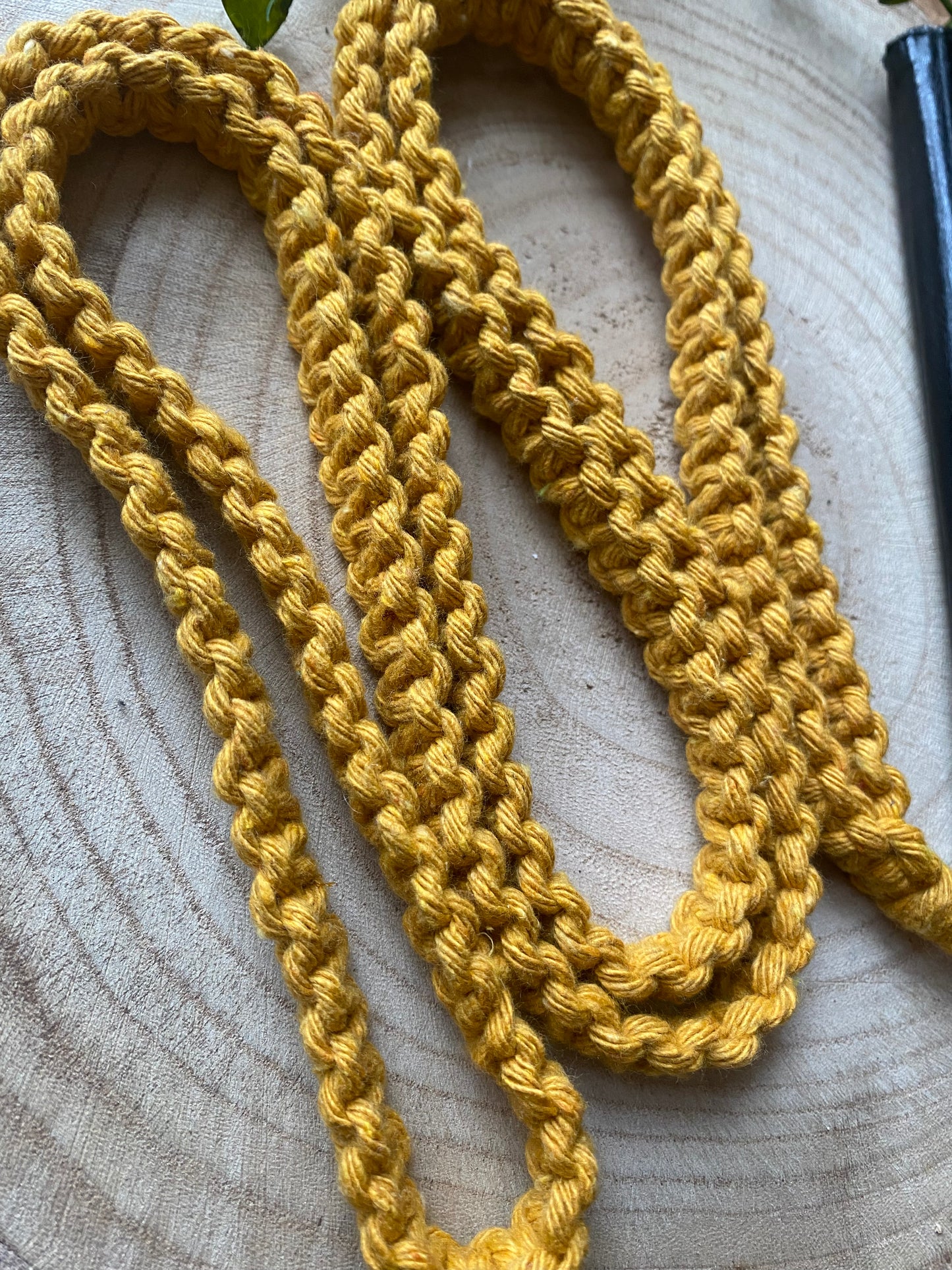A close-up of a Macra-Made-With-Love Recycled Cotton Crossbody Phone Strap arranged in multiple layers on a light wooden surface. The texture and stitch patterns of the eco-friendly crochet work are prominently displayed. A small portion of a green leaf is visible in the upper left corner.