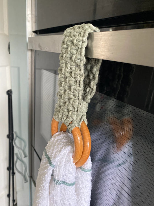 A handmade macrame kitchen tea towel ring holder, made with sage green cotton yarn, and two wooden rings, is looped over a silver oven handle, and holds a white tea towel with green stripes between the wooden rings.