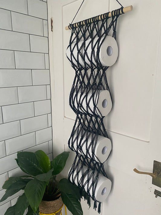 8 white toilet rolls, held securely in a black knotted macrame toilet roll holder, on a long wooden dowel, hooked on to the back of a white door next to a green houseplant.