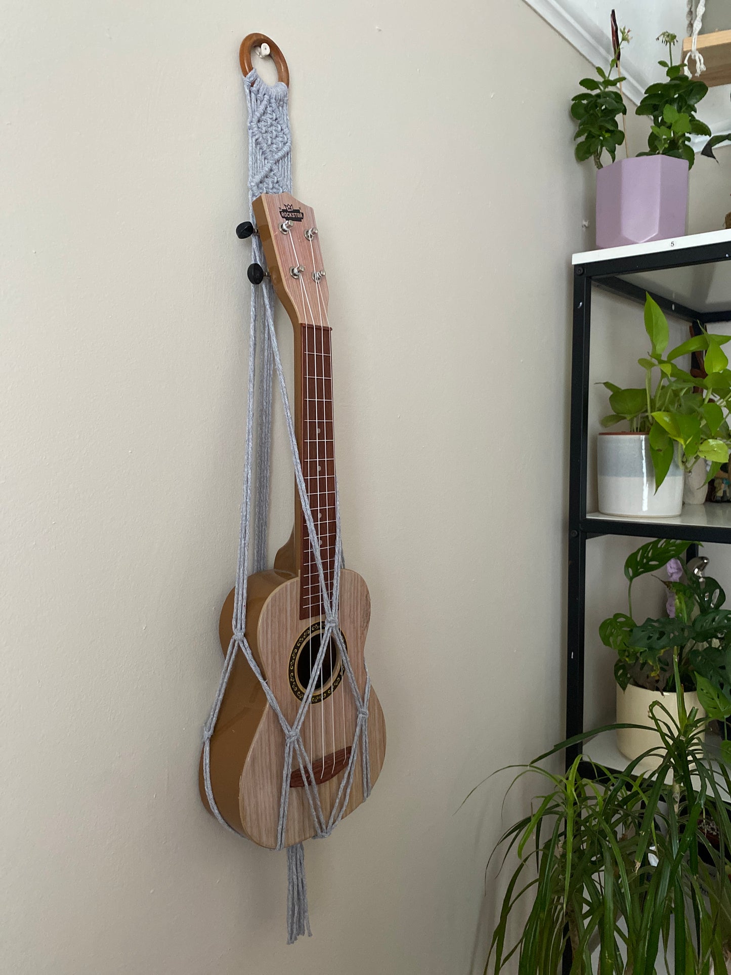 A light brown ukulele sits inside of a handmade macrame wall hanging ukulele hanger, which is made with light grey recycled cotton, and a repurposed wooden hanging ring. A tall shelving unit full of green leafy houseplants is to the right of the image.