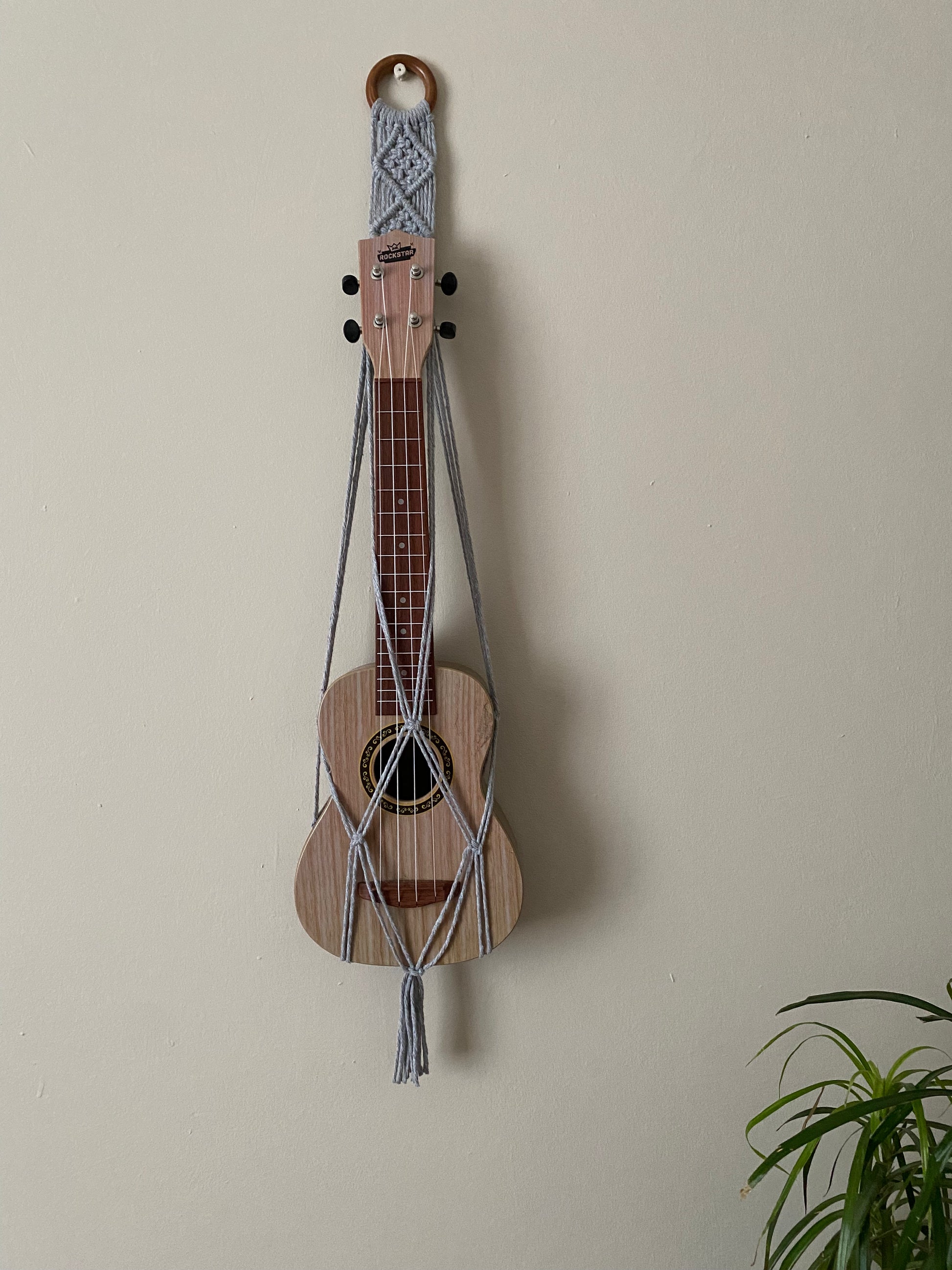 A light brown ukulele sits inside of a handmade macrame wall hanging ukulele hanger, which is made with light grey recycled cotton, and a repurposed wooden hanging ring.