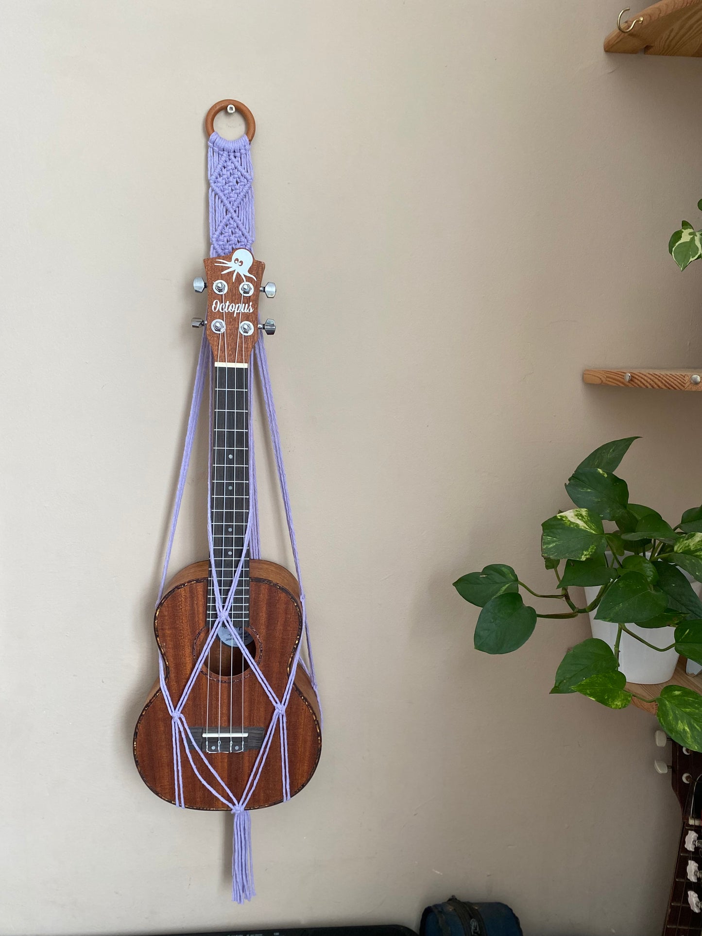A dark brown ukulele sits inside of a handmade macrame wall hanging ukulele hanger, which is made with lilac recycled cotton, and a repurposed wooden hanging ring. A large green leafy plant is to the right of the image.