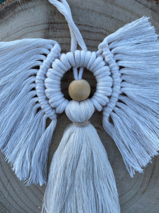 Handmade macrame hanging angel decoration with white cords and wooden bead, displayed against a wooden background.
