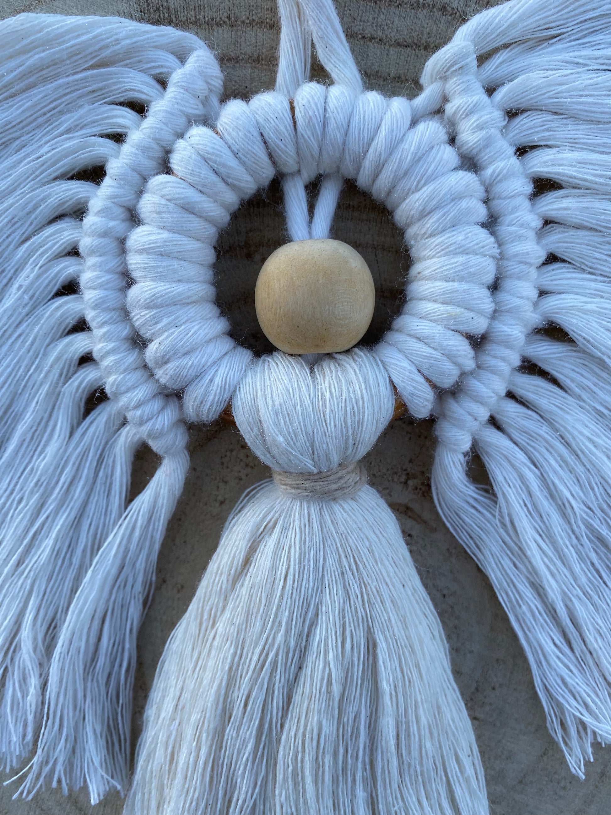 Close up of a handmade macrame hanging angel decoration with white cords and wooden bead, displayed against a wooden background.