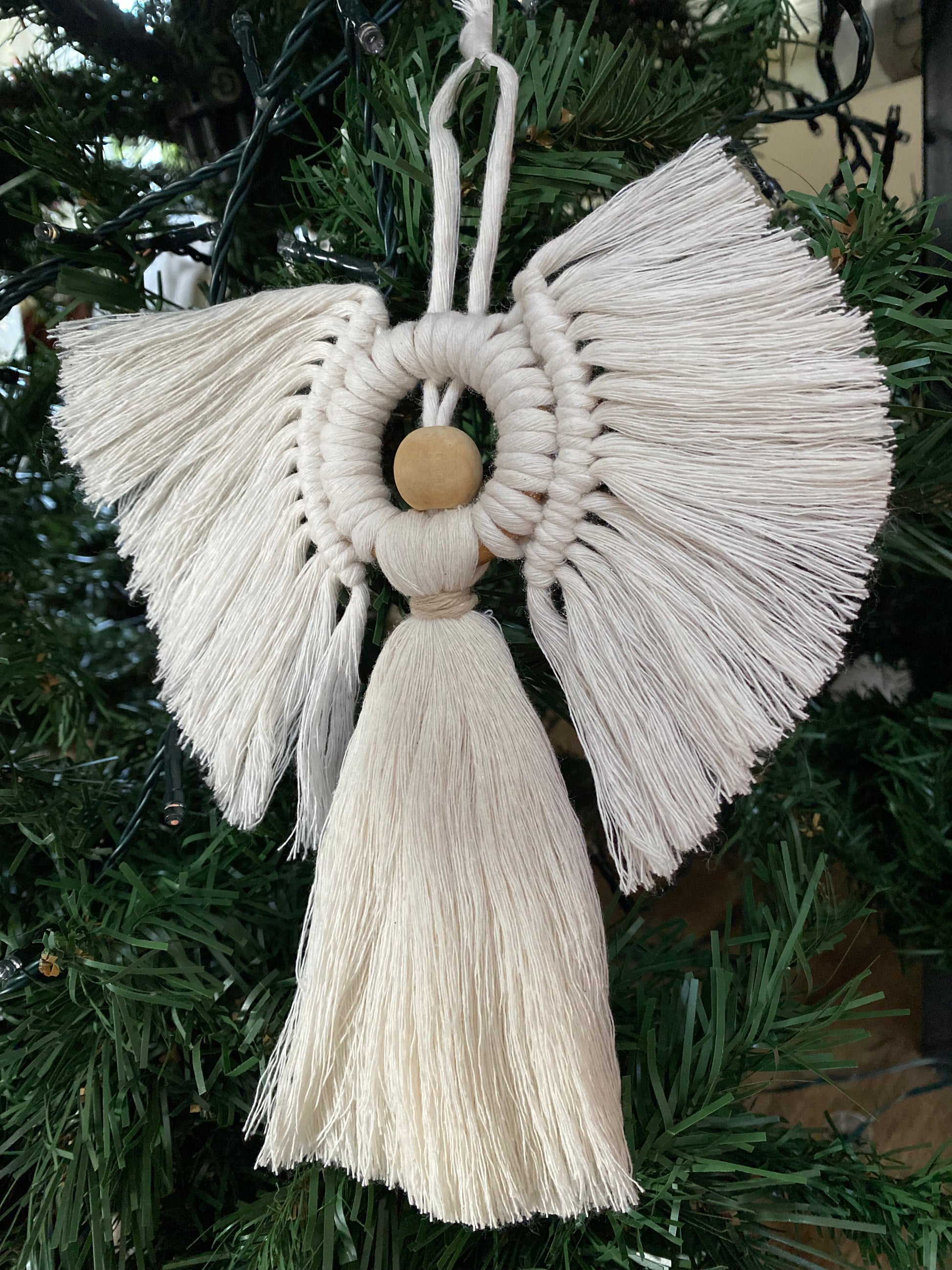 Handmade macrame hanging angel decoration with white cords and wooden bead, displayed hanging in a dark green artificial christmas tree.
