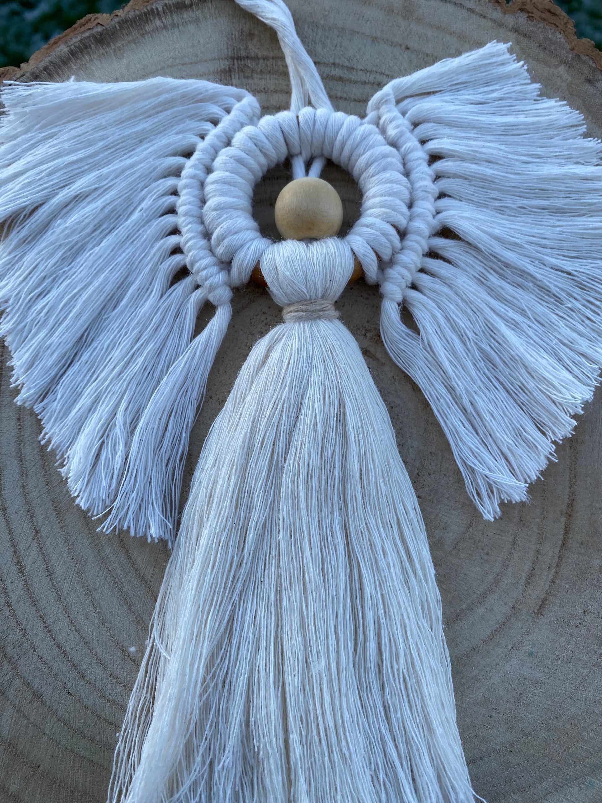 Angled view of a handmade macrame hanging angel decoration with white cords and wooden bead, displayed against a wooden background.