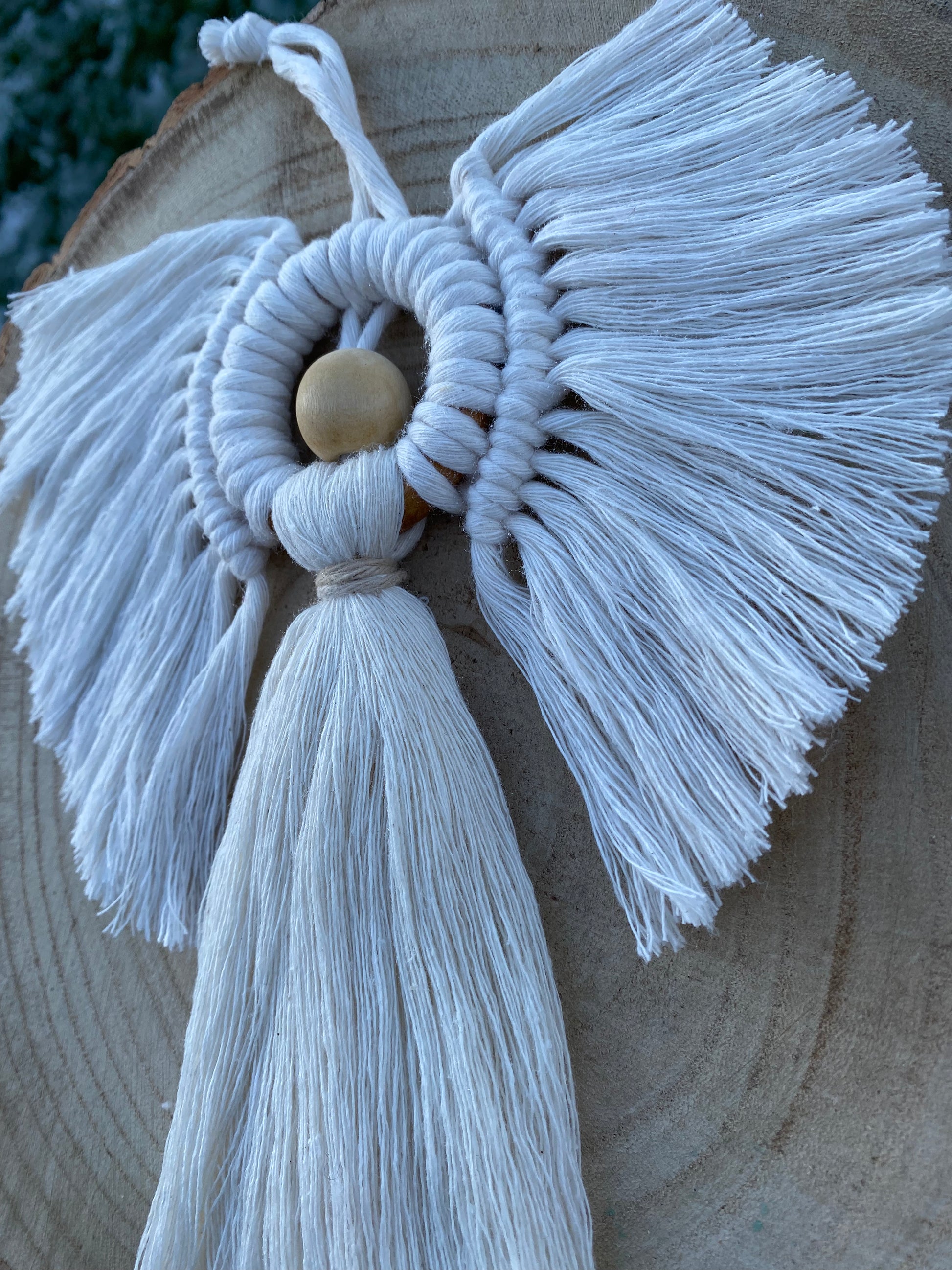 Side view of a handmade macrame hanging angel decoration with white cords and wooden bead, displayed against a wooden background.