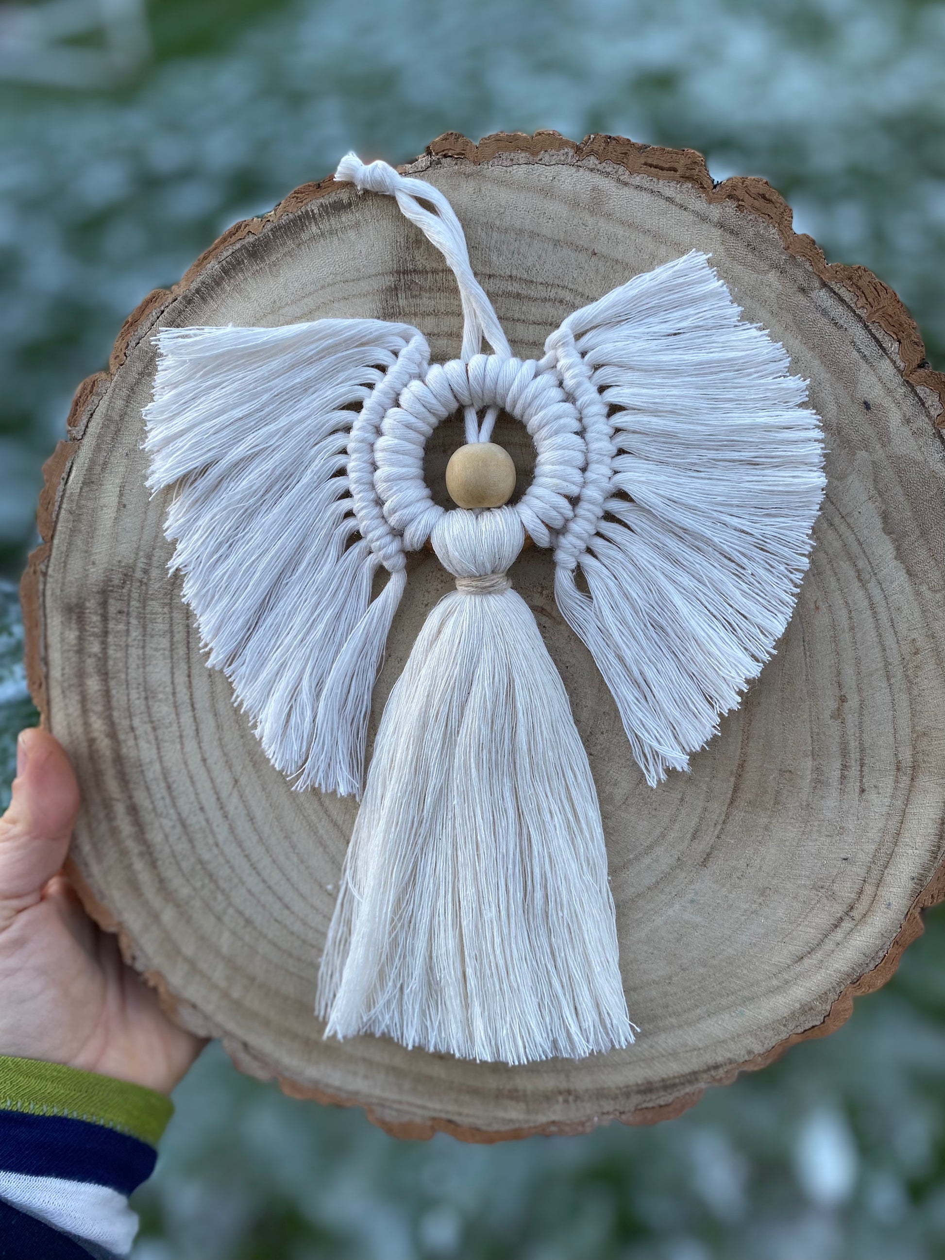Handmade macrame hanging angel decoration with white cords and wooden bead, displayed on a wooden board, held outside with frosty grass blurred in the background.