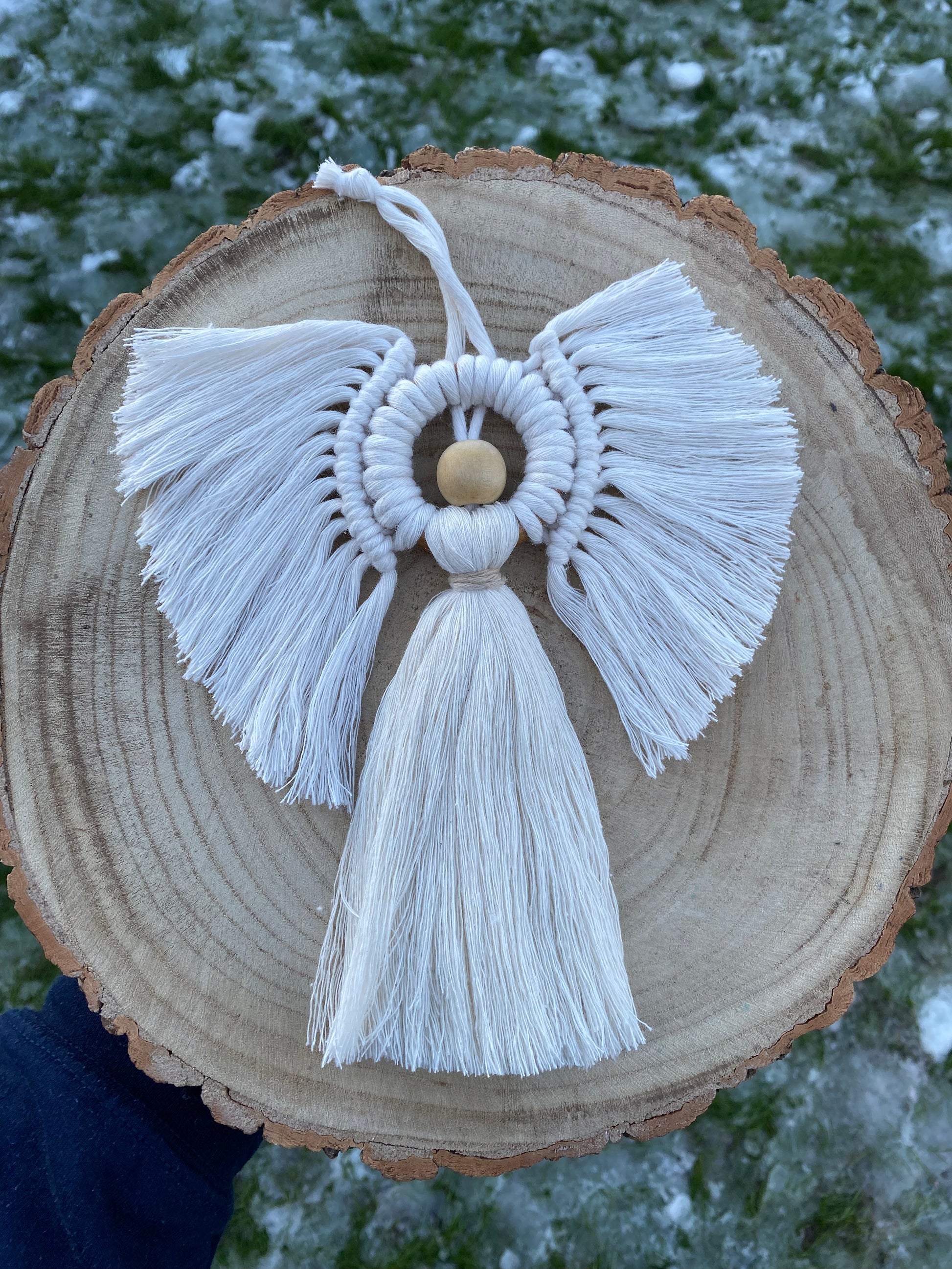 Handmade macrame hanging angel decoration with white cords and wooden bead, displayed on a log slice with frosty grass underneath.