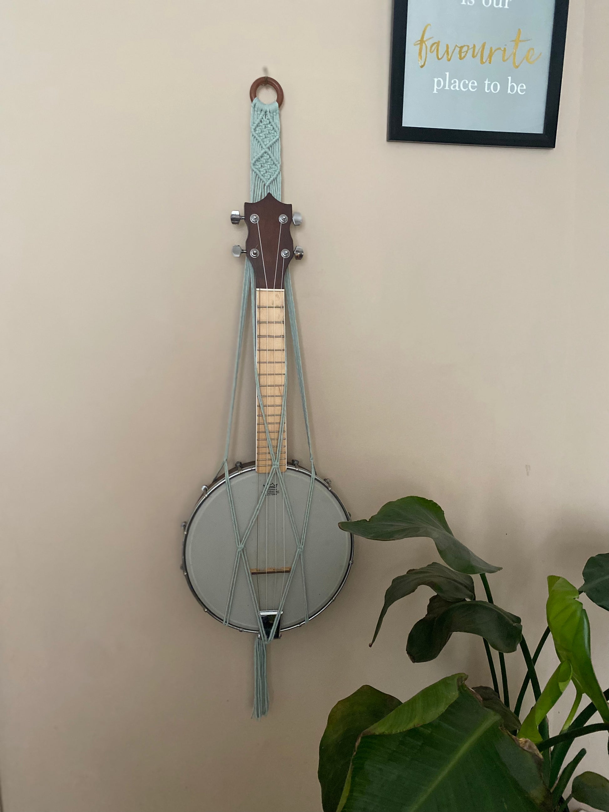 A banjolele instrument sits securely in a macrame wall mounted hanger, made with pastel green coloured yarn and a wooden hanging ring. Displayed against a beige painted wall, with a leafy houseplant nearby.
