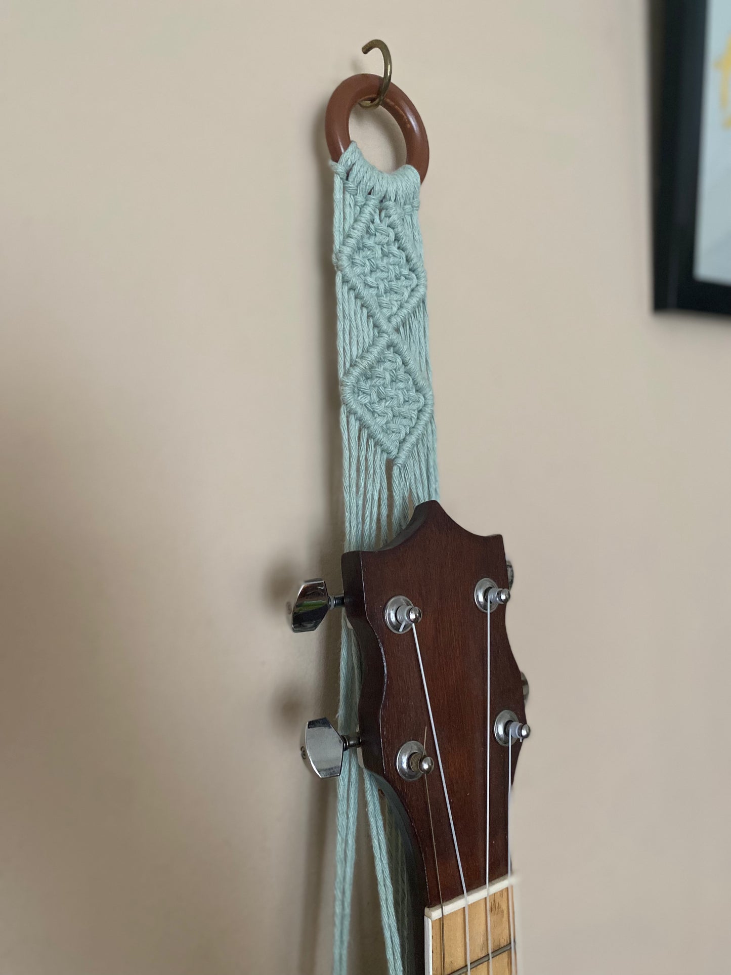 A close up image of the top section of a banjolele instrument held in a macrame wall mounted hanger, made with pastel green coloured yarn and a wooden hanging ring. Displayed against a beige painted wall.