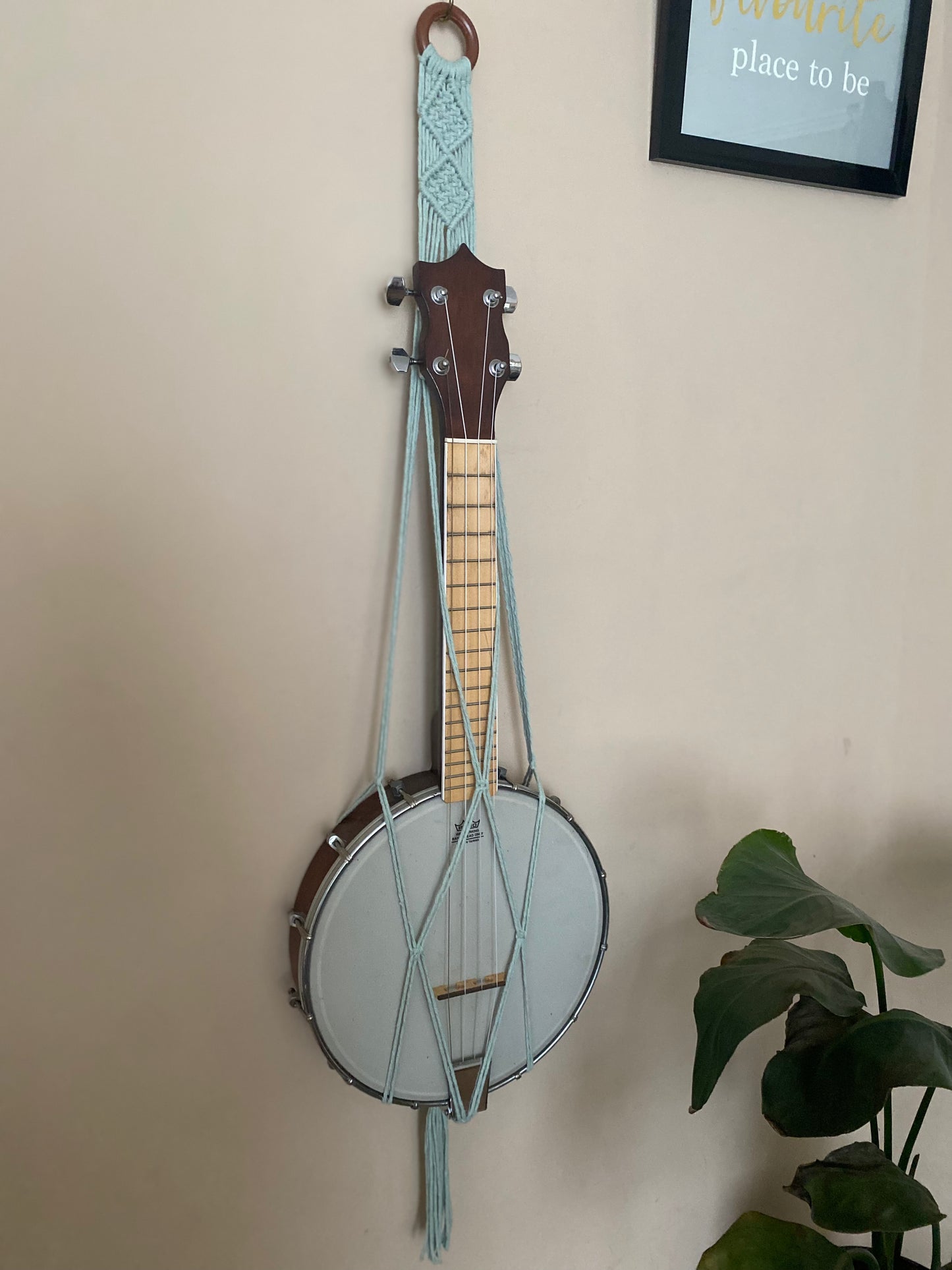 A banjolele instrument sits securely in a macrame wall mounted hanger, made with pastel green coloured yarn and a wooden hanging ring. Displayed against a beige painted wall, with a large houseplant nearby.