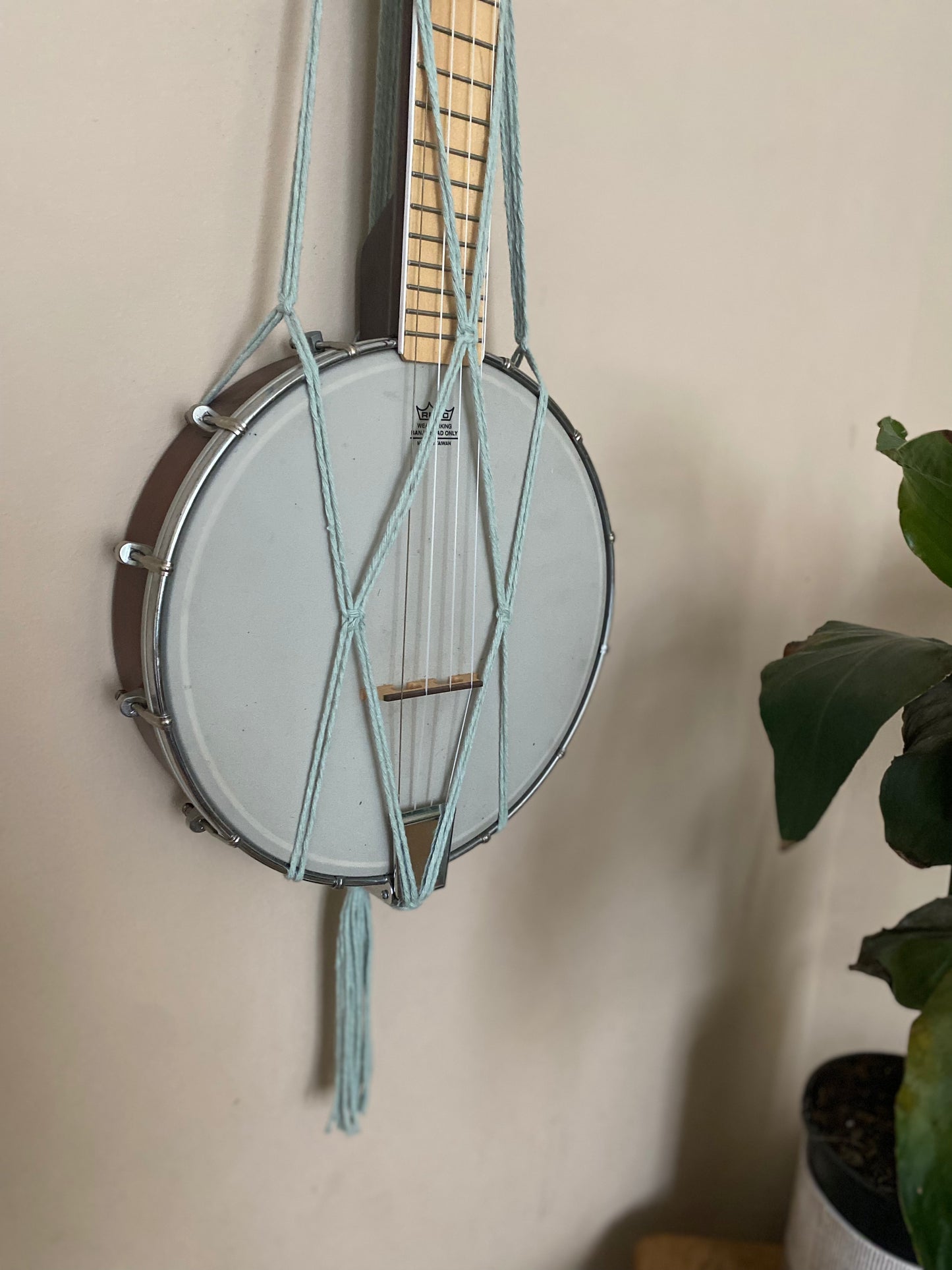 Close up image of the bottom part of a banjolele instrument held in a macrame wall mounted hanger, made with pastel green coloured yarn. Displayed against a beige painted wall.