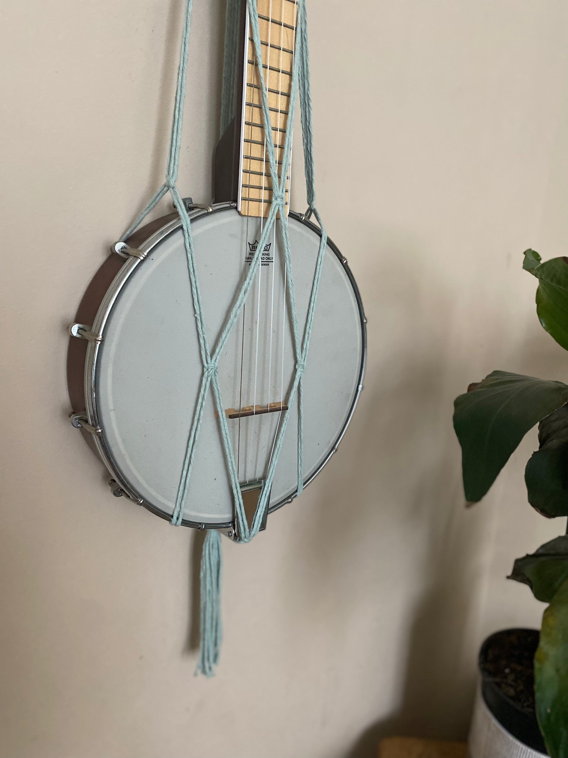Close up image of the bottom part of a banjolele instrument held in a macrame wall mounted hanger, made with pastel green coloured yarn. Displayed against a beige painted wall.