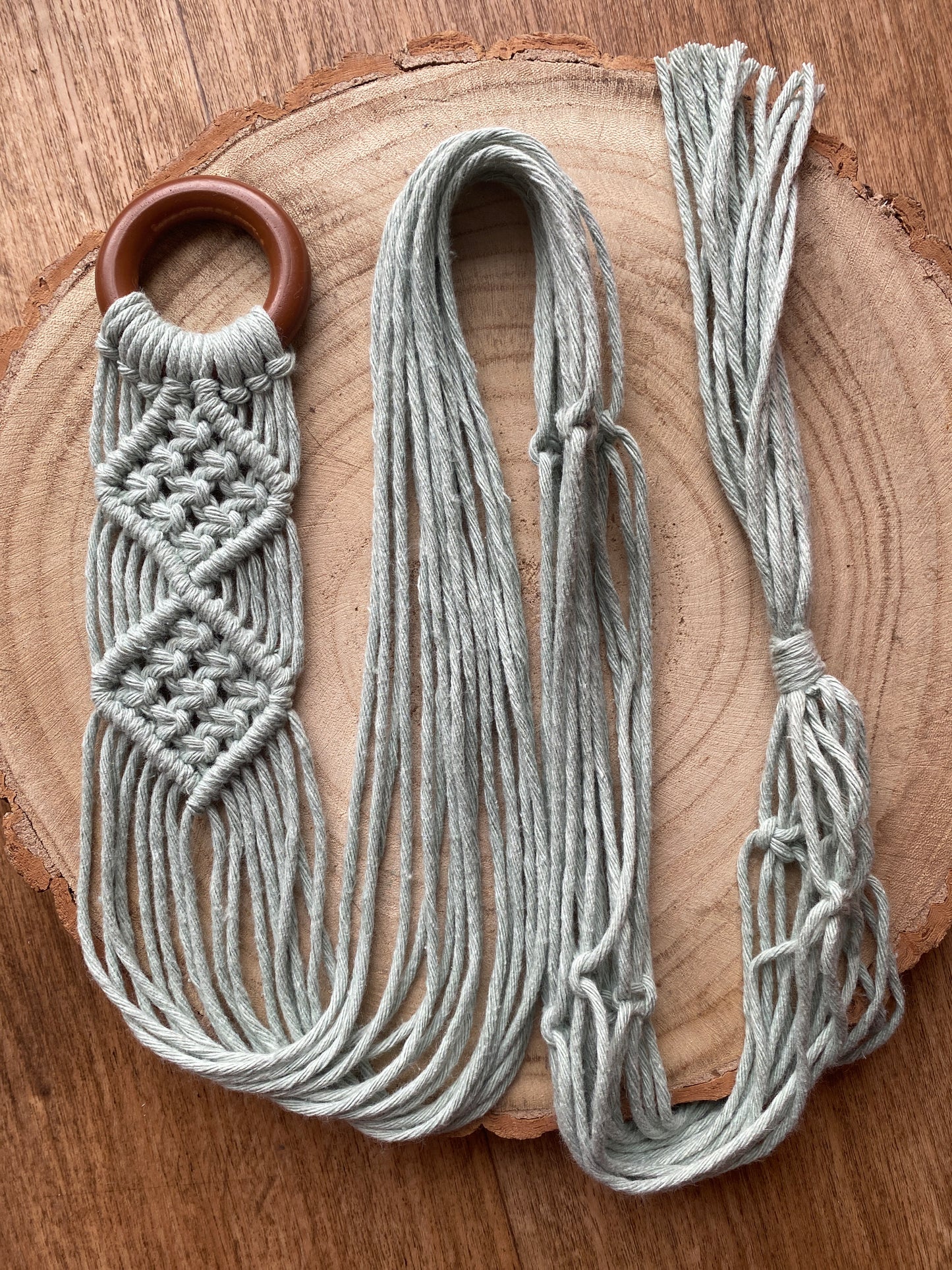 A handmade banjolele macrame instrument hanger, made with pastel green recycled cotton on a dark wooden ring, is neatly laid out across a circular wooden surface, showcasing an intricate knot pattern.