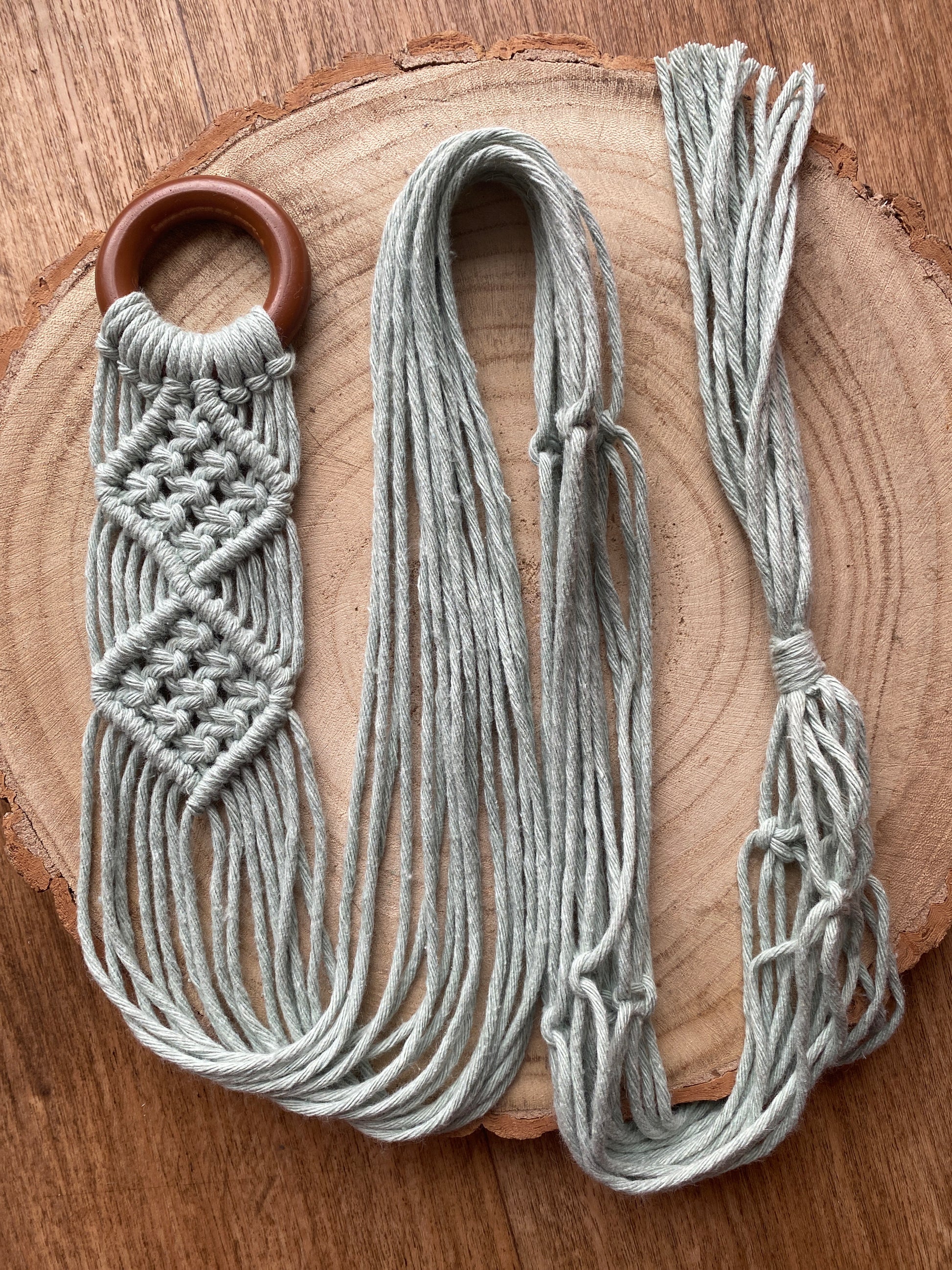 A handmade banjolele macrame instrument hanger, made with pastel green recycled cotton on a dark wooden ring, is neatly laid out across a circular wooden surface, showcasing an intricate knot pattern.