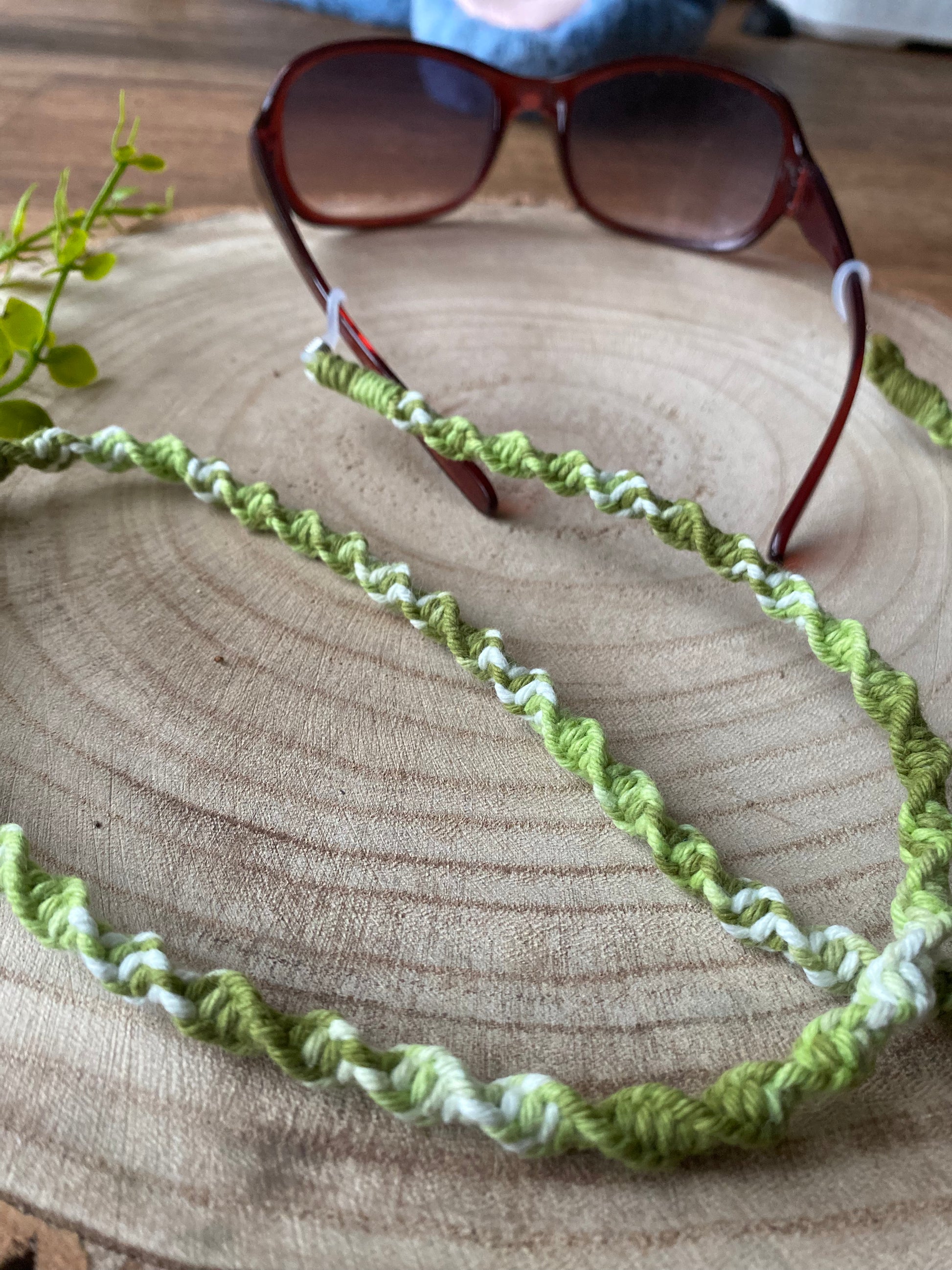 A pair of dark sunglasses are placed on a wooden surface with a handmade macrame eyewear holder chain, made with variegated green recycled cotton yarn, attached to them.