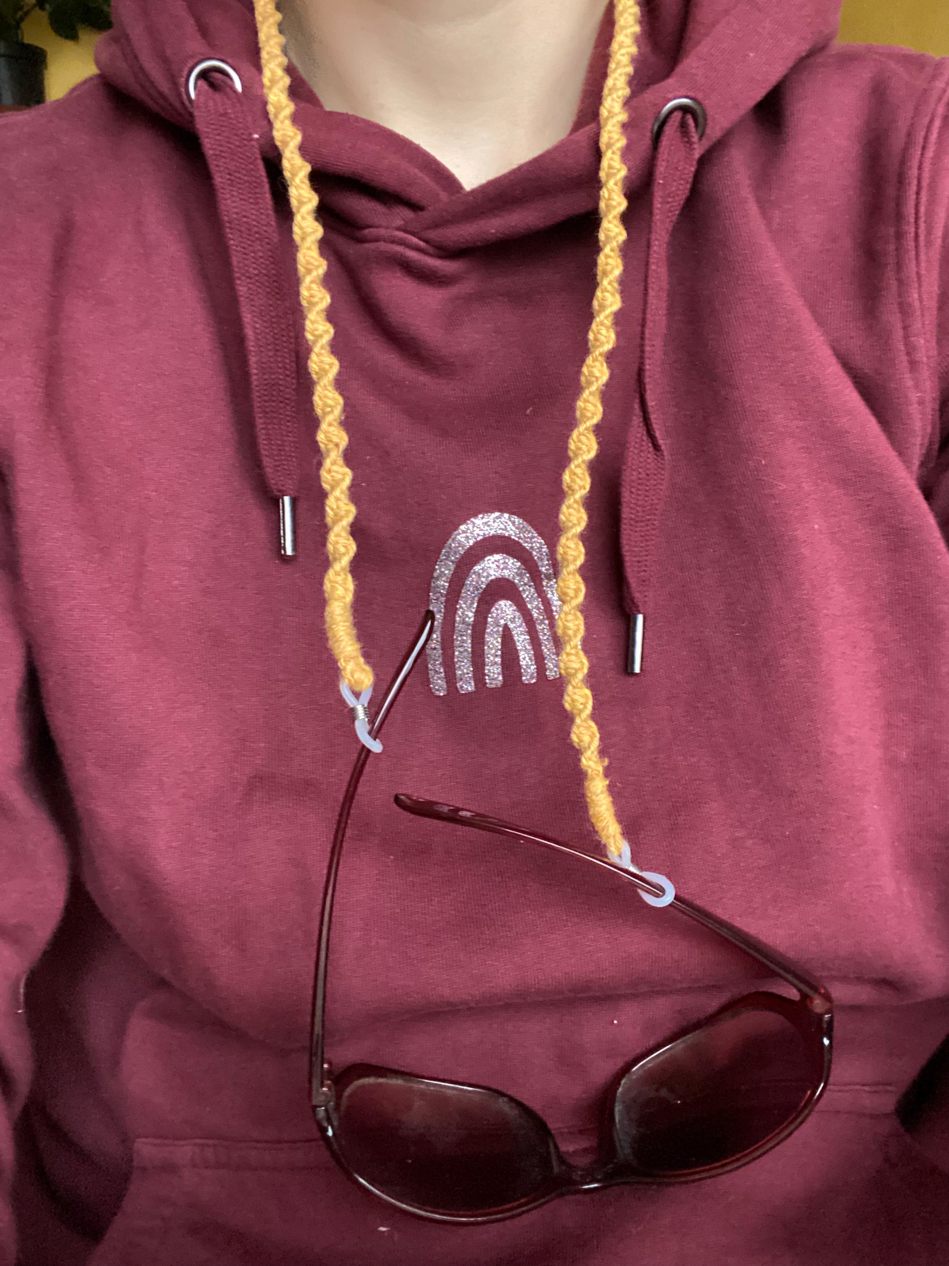 A woman wearing a burgundy hoodie, has a pair of dark sunglasses attached to a handmade macrame eyewear holder chain, made with mustard yellow recycled cotton yarn, around her neck.