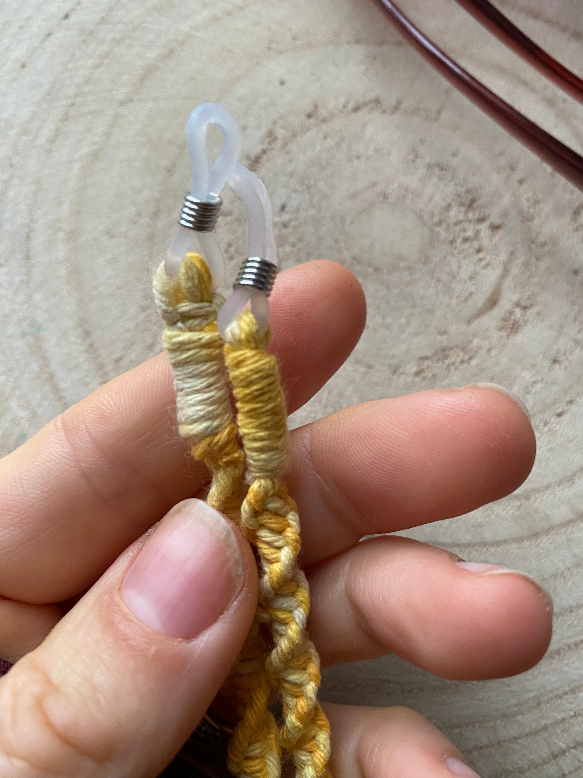 A hand shows clear loops on the ends of a handmade macrame eyewear holder chain, made with variegated yellow recycled cotton yarn.