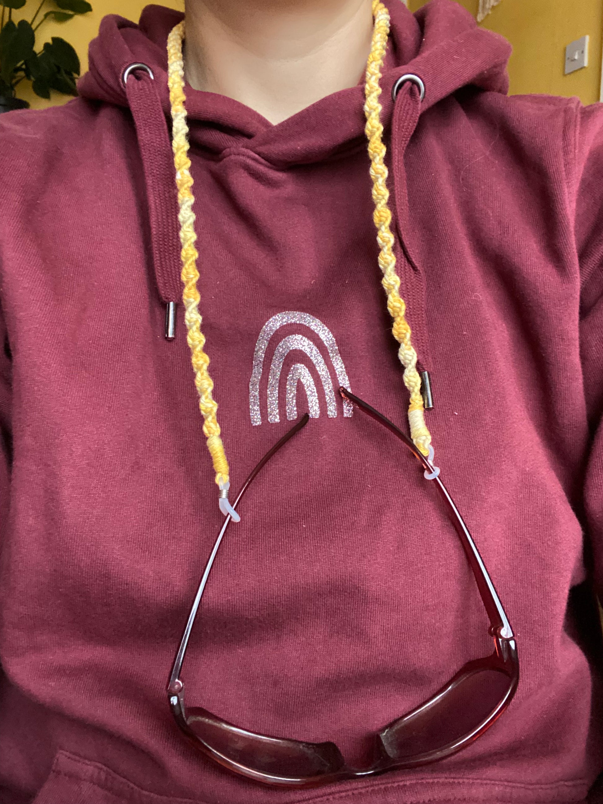 A woman in a burgundy hoodie, wears a pair of dark sunglasses attached to a handmade macrame eyewear holder chain, made with variegated yellow recycled cotton yarn, around her neck.