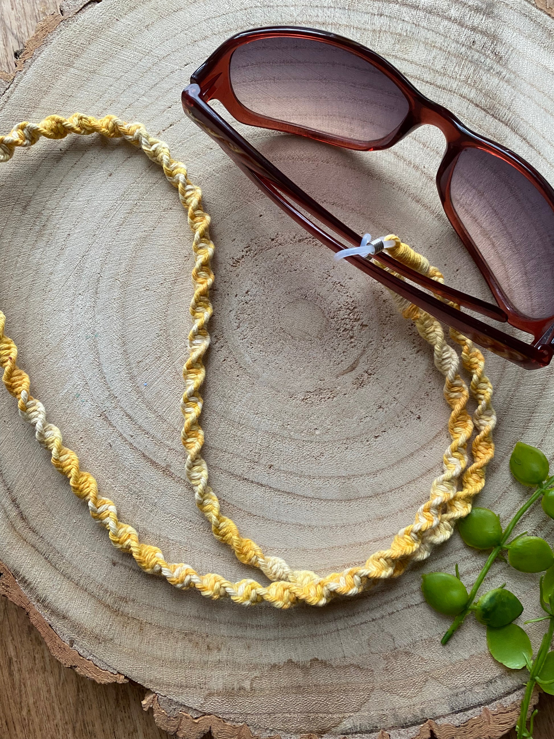 A pair of dark sunglasses are placed on a wooden surface with a handmade macrame eyewear holder chain, made with variegated yellow recycled cotton yarn, attached to them.
