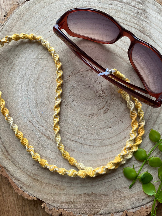 A pair of dark sunglasses are placed on a wooden surface with a handmade macrame eyewear holder chain, made with variegated yellow recycled cotton yarn, attached to them.