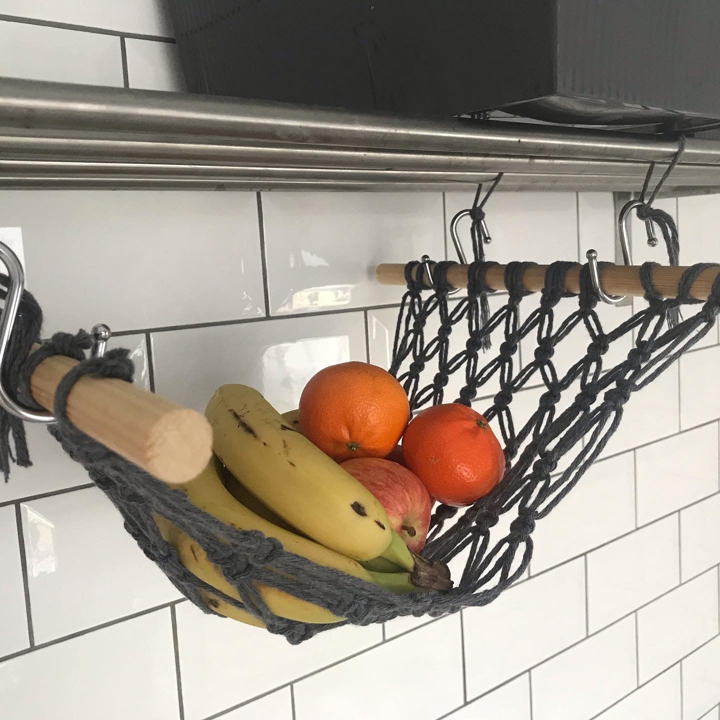 Bananas, oranges and apples hang in a macrame hammock made with dark grey recycled cotton and wooden dowels, underneath a metal shelf on a white tiled wall.