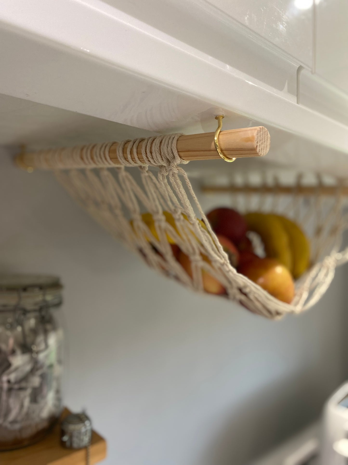 Close up image of a macrame fruit hammock made with natural recycled cotton and wooden dowels, hanging underneath a white kitchen wall unit on gold hooks.