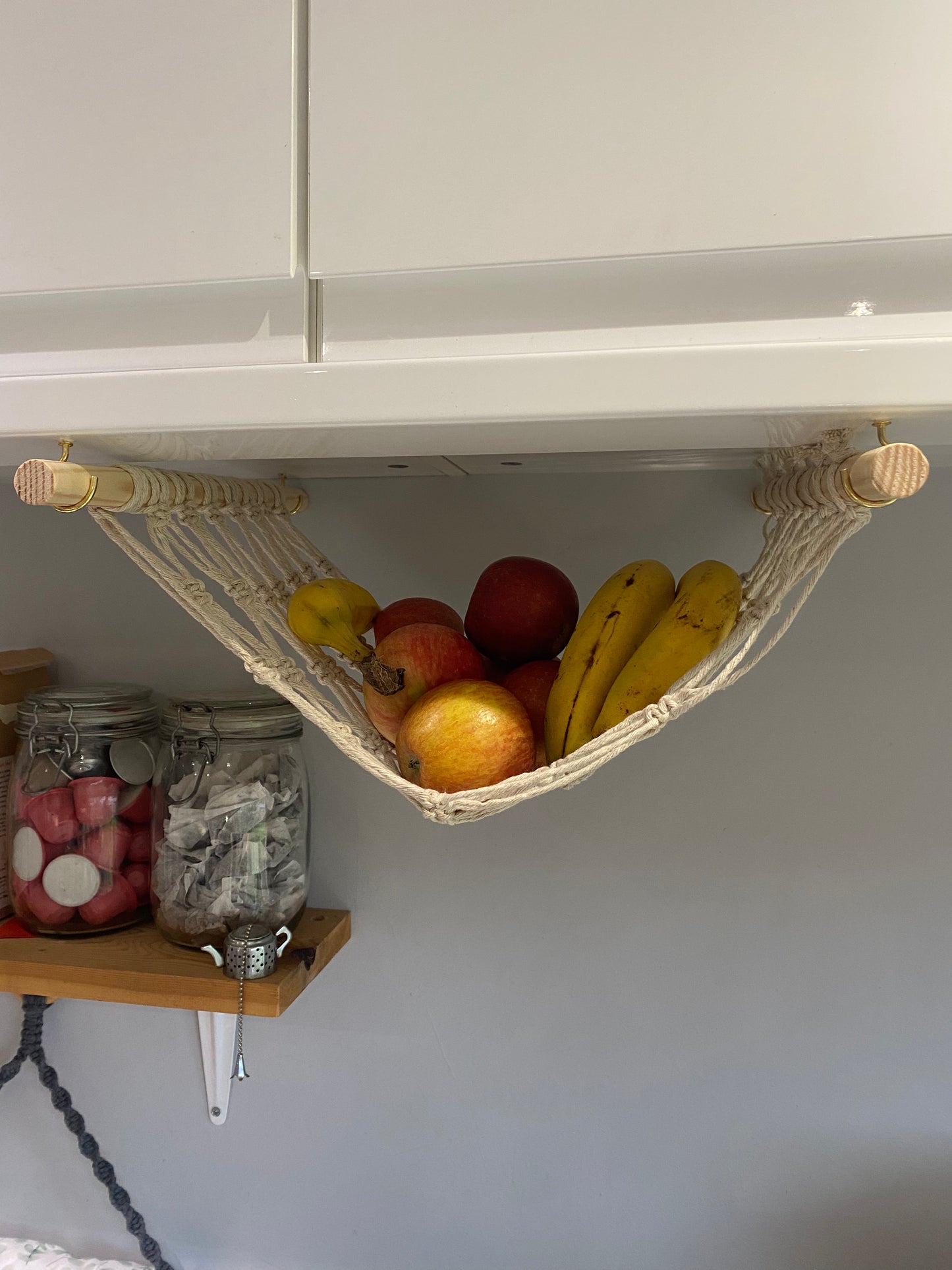 Bananas and apples hang in a macrame hammock made with natural recycled cotton and wooden dowels, underneath a white kitchen wall unit. There are glass jars with tea bags and coffee pods inside on a wooden shelf in the background.