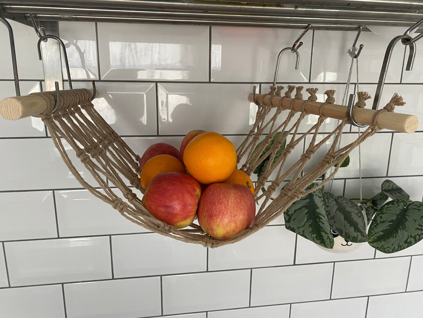 Oranges and apples hang in a macrame hammock made with taupe recycled cotton and wooden dowels, underneath a metal wall shelf using metal S hooks.