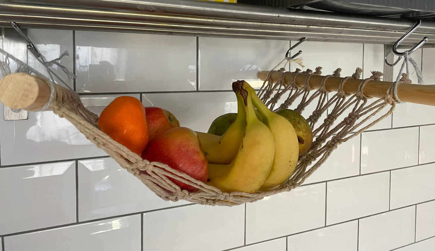 Close up of bananas, oranges and apples hanging in a macrame hammock made with taupe recycled cotton and wooden dowels, underneath a metal wall shelf.