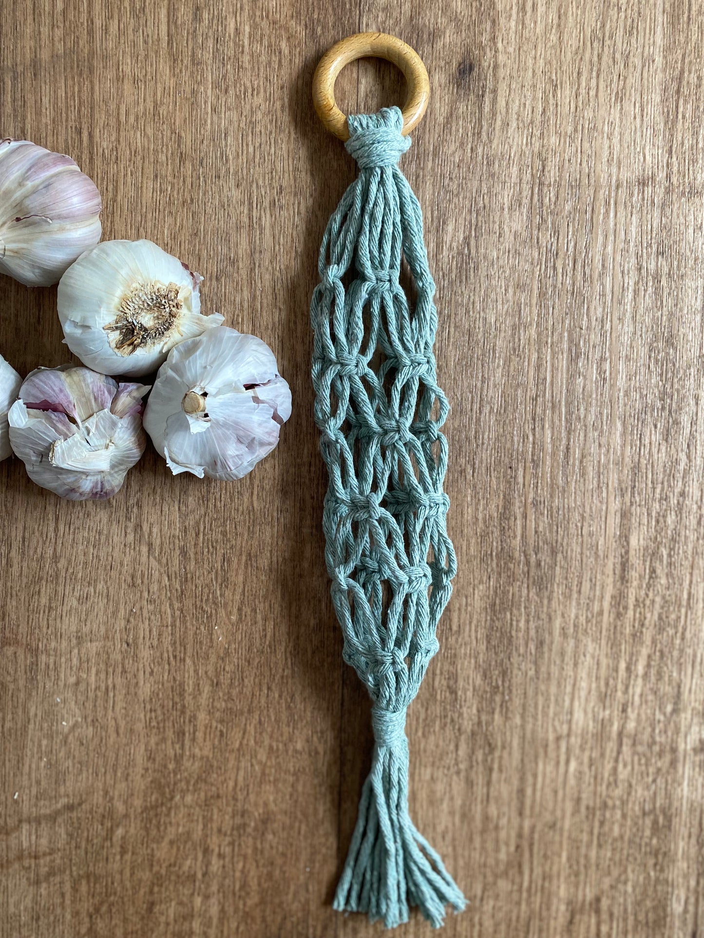 A handmade macrame garlic hanger made with green cotton yarn and a wooden ring, laid on a wooden floor next to a pile of fresh garlic bulbs.