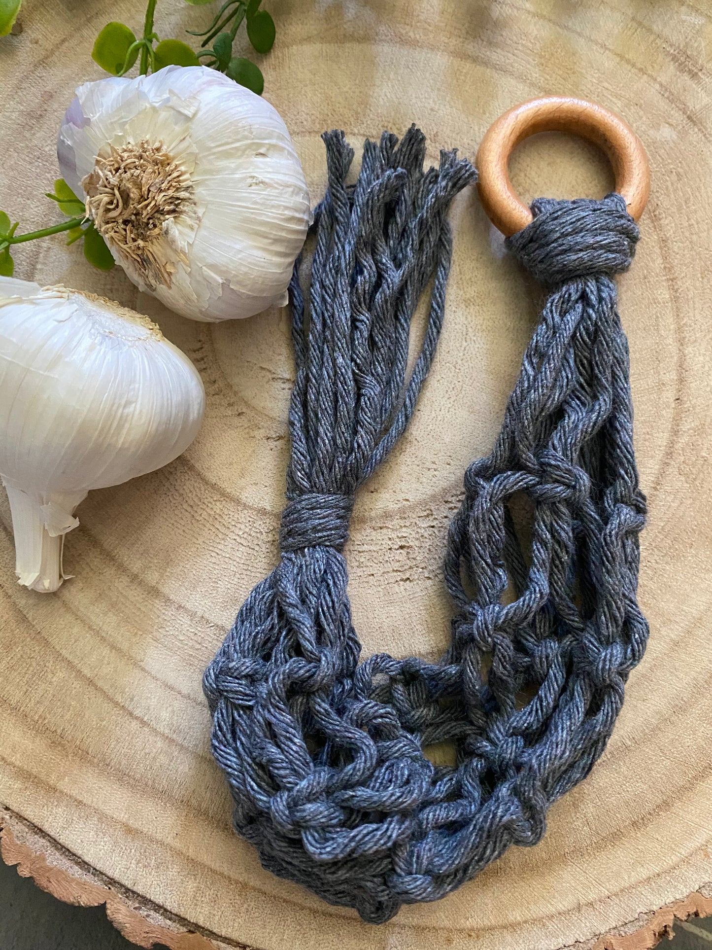 A handmade macrame garlic hanger made with dark grey cotton yarn and a wooden ring, laid on a round wooden board next to two fresh garlic bulbs.