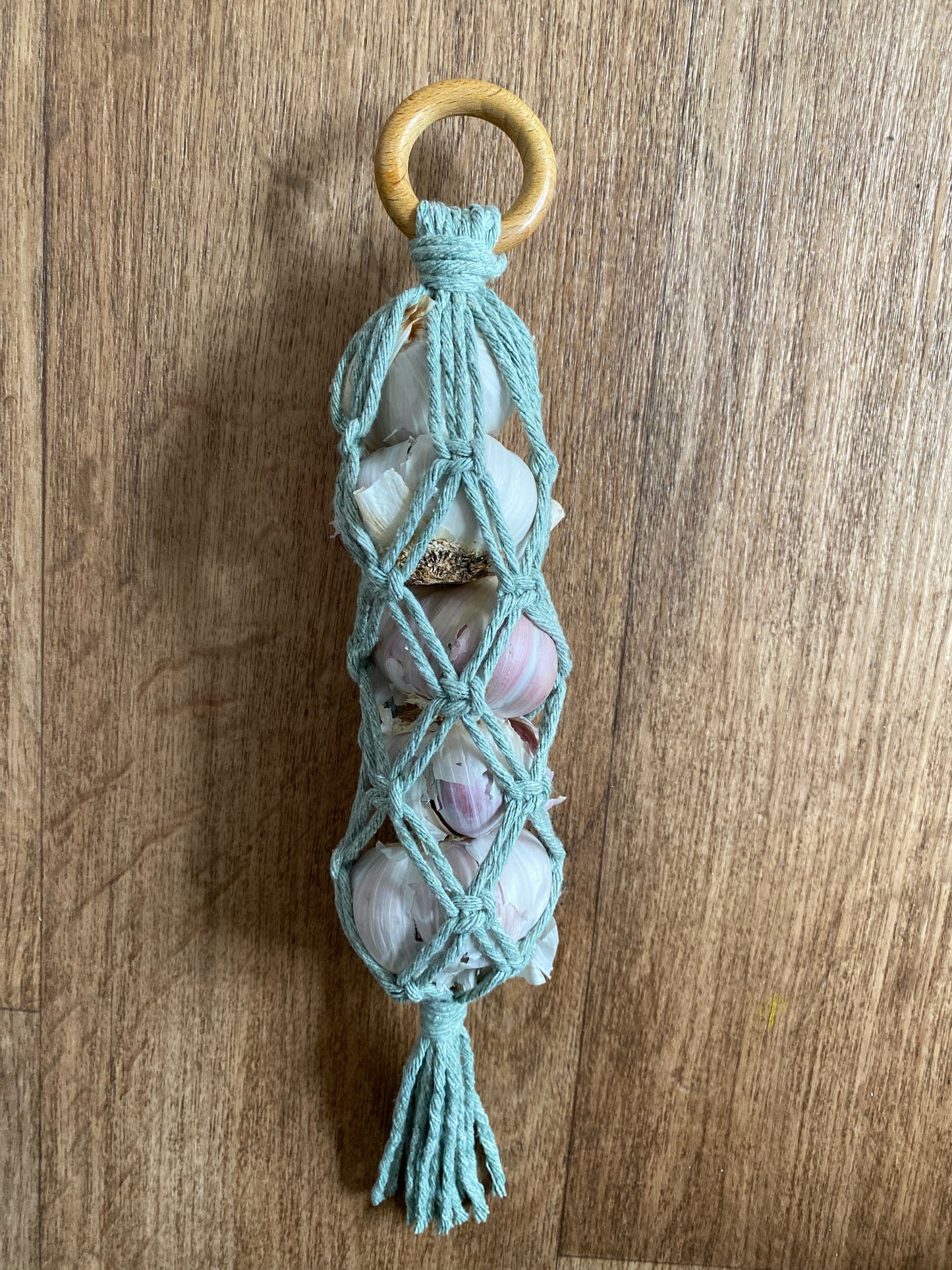 A handmade macrame garlic hanger made with green cotton yarn and a wooden ring, laid on a wooden floor holding 5 fresh garlic bulbs neatly inside the macramé knots.