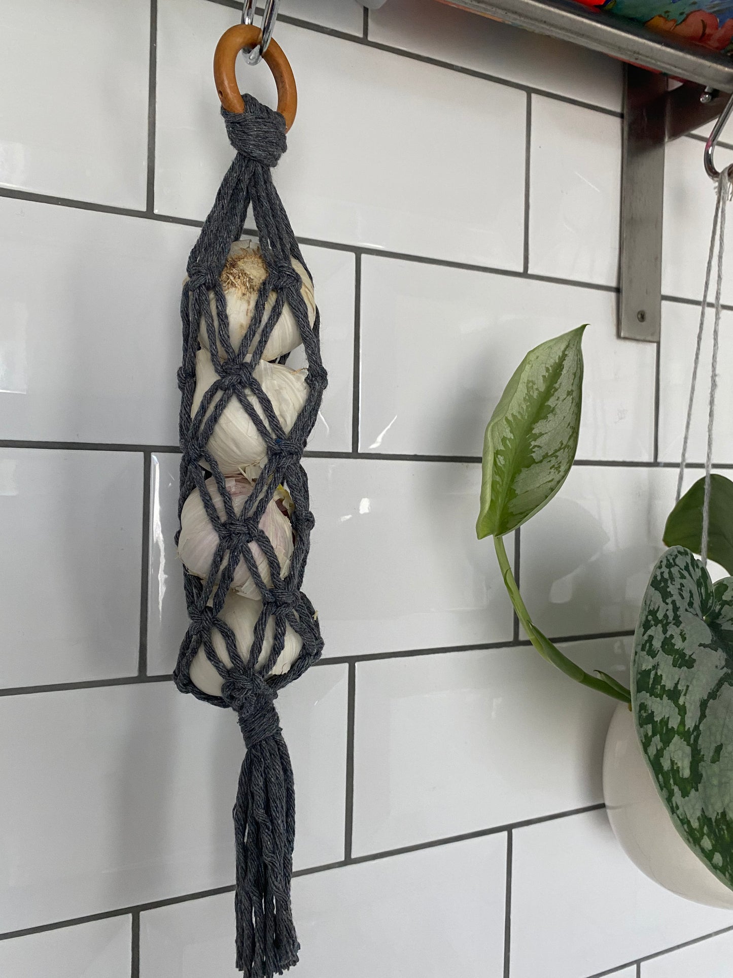 A handmade macrame garlic hanger made with dark grey cotton yarn and a wooden ring, holding 4 fresh garlic bulbs, hanging against a white tiled wall.