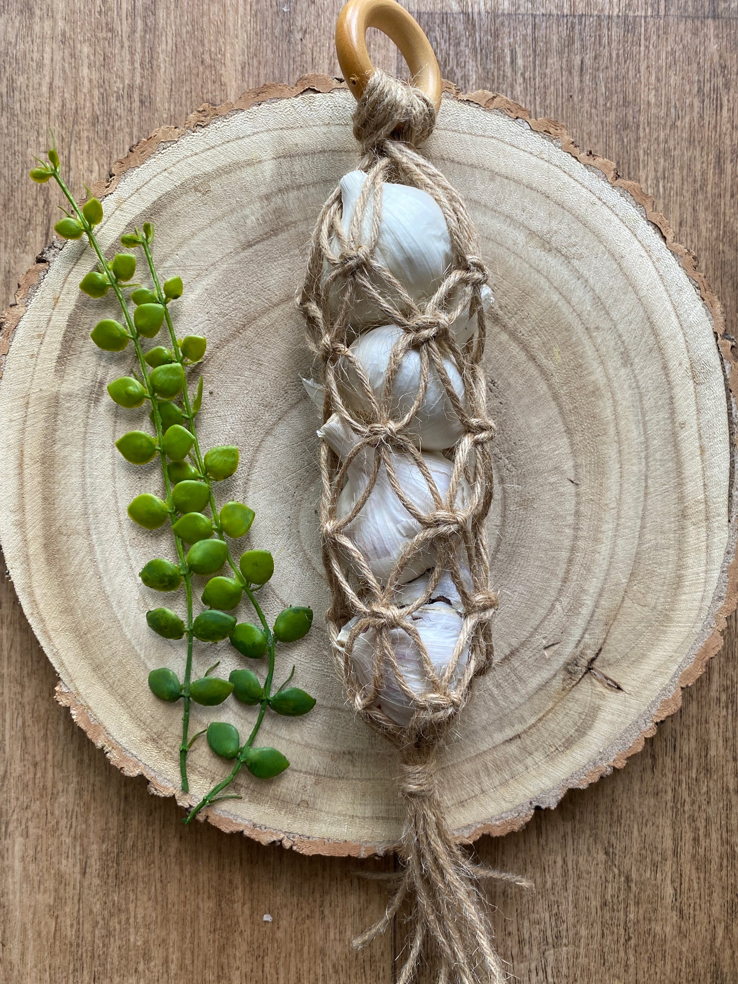 A handmade macrame garlic hanger made with jute twine and a wooden ring, laid on a wooden board with 4 fresh garlic bulbs inside.
