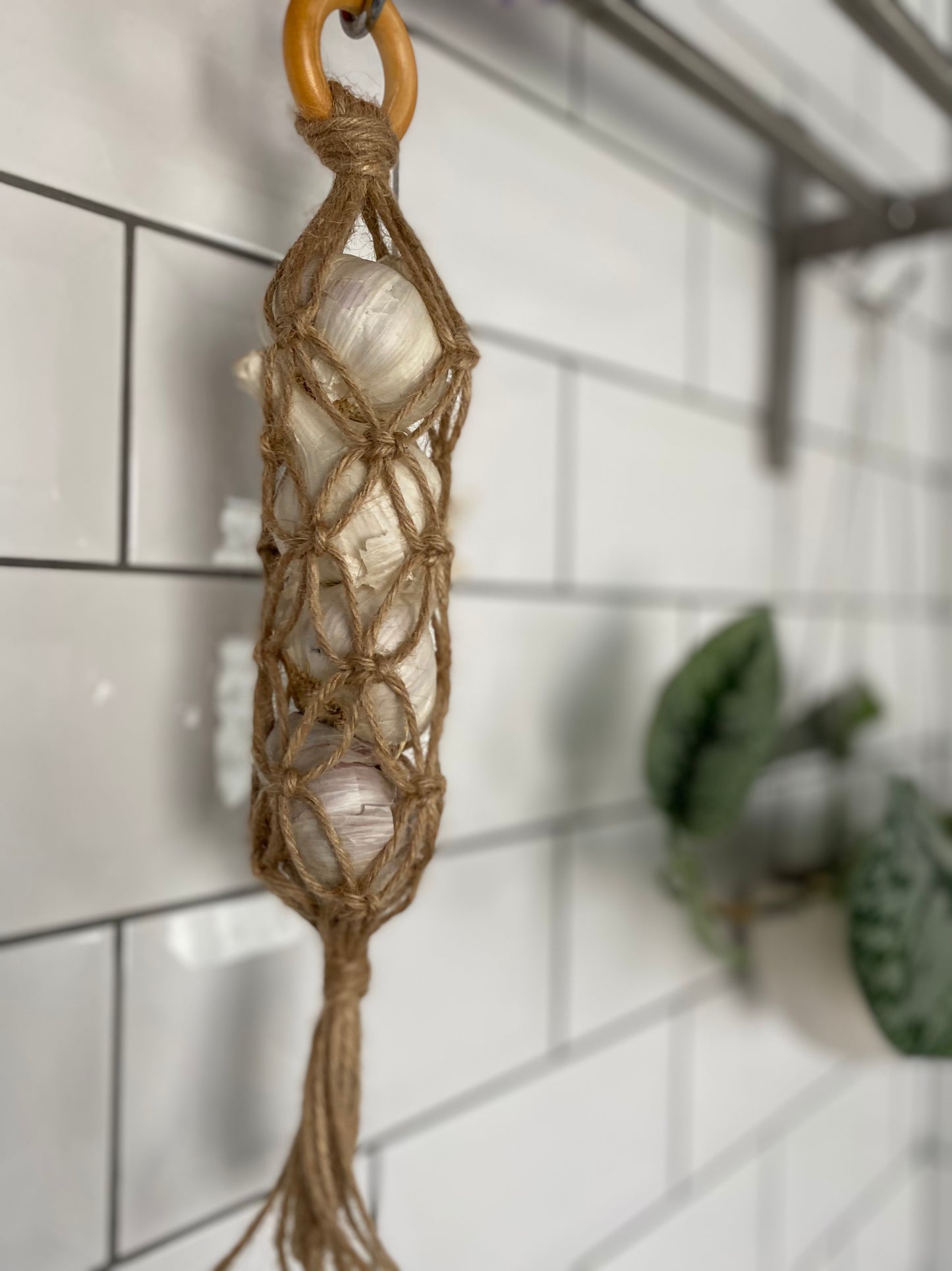 A handmade macrame garlic hanger made with jute twine and a wooden ring, holding 4 fresh garlic bulbs, hanging against a white tiled wall.