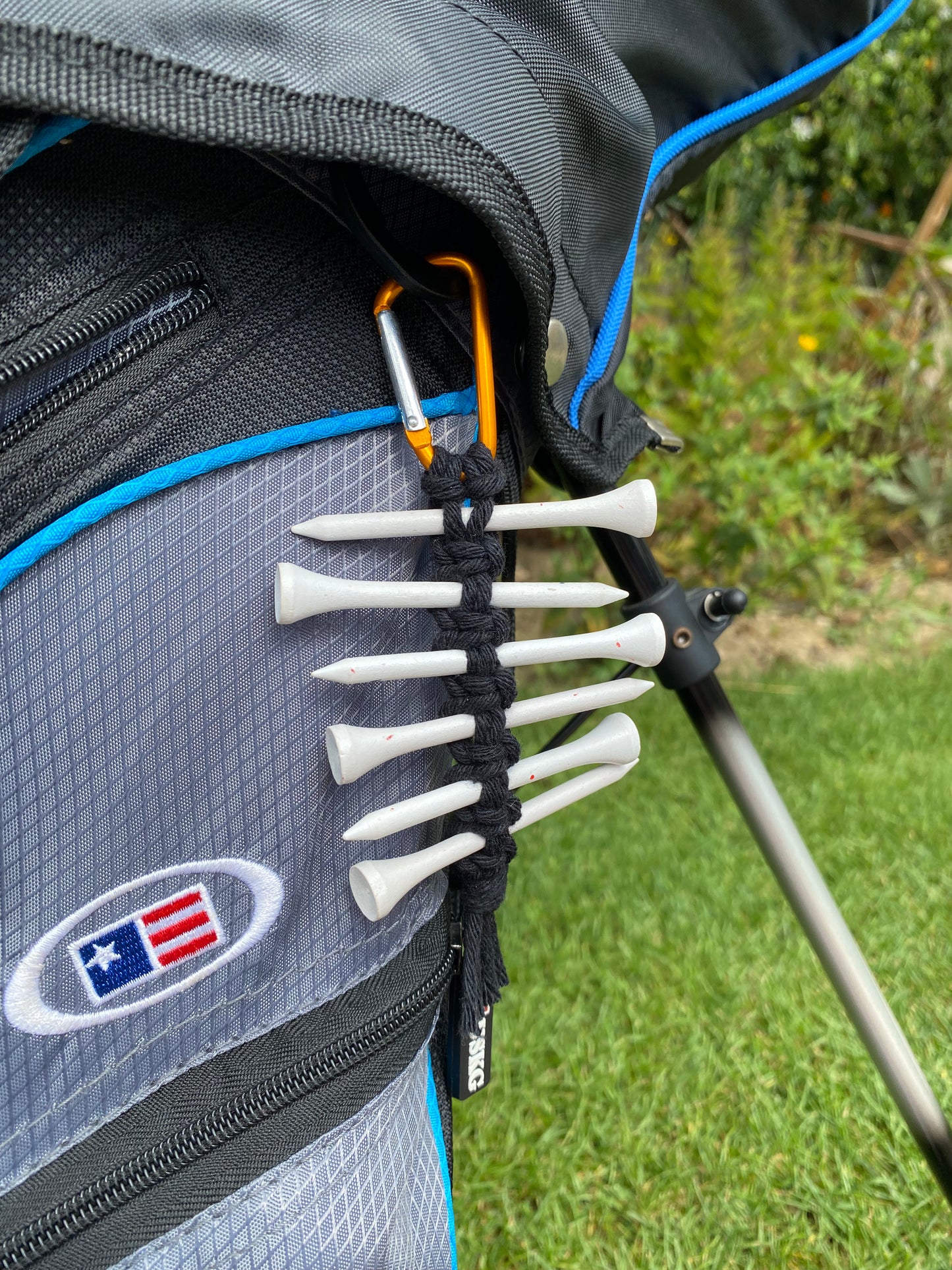 A knotted golf tee holder, made with black yarn on an orange gold carabiner clip, attached on to the side of a black golf bag, holding 6 white golf tees.