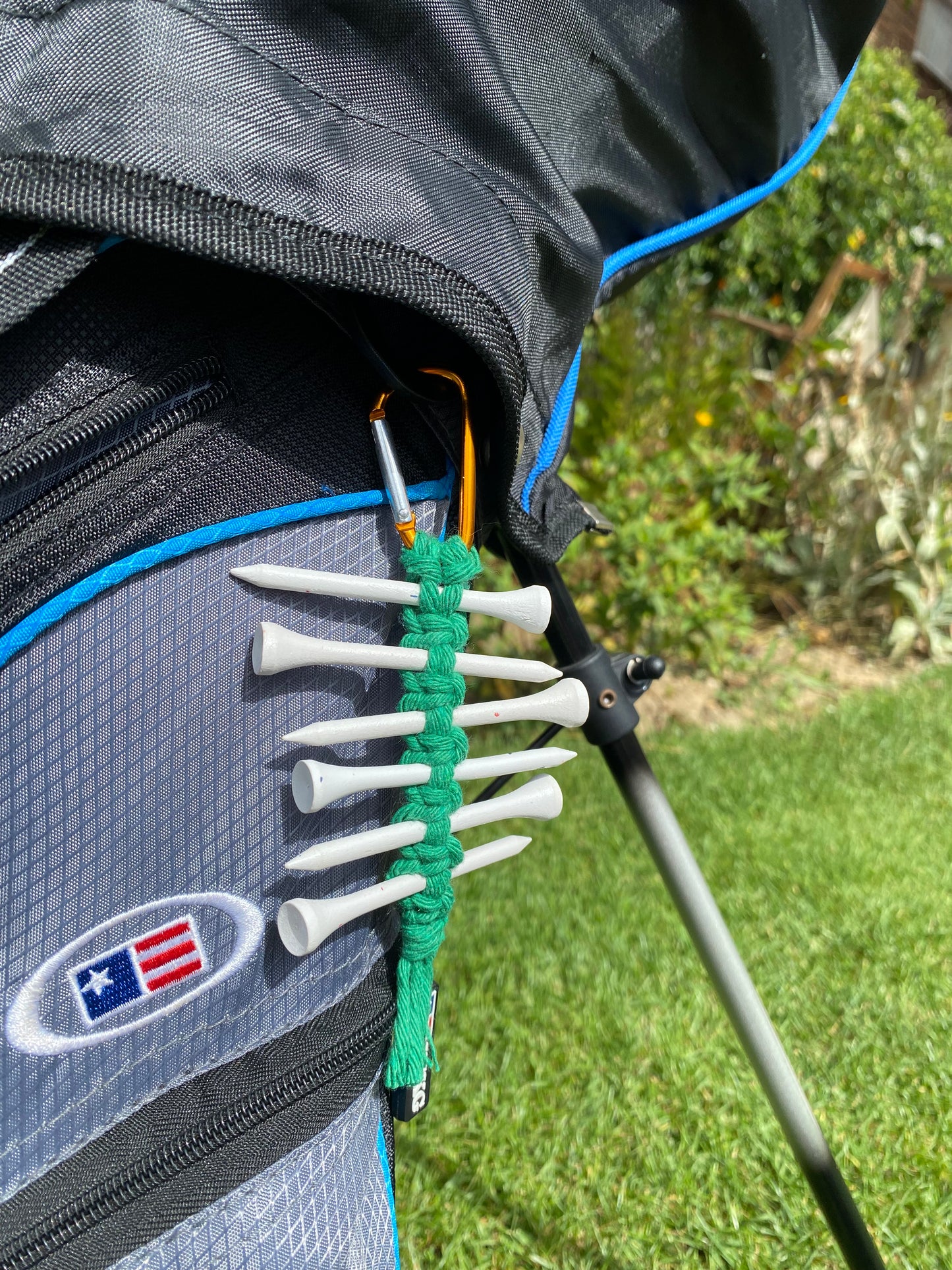 A knotted golf tee holder, made with green yarn on an orange gold carabiner clip, attached to a black golf bag holding 6 white golf tees.